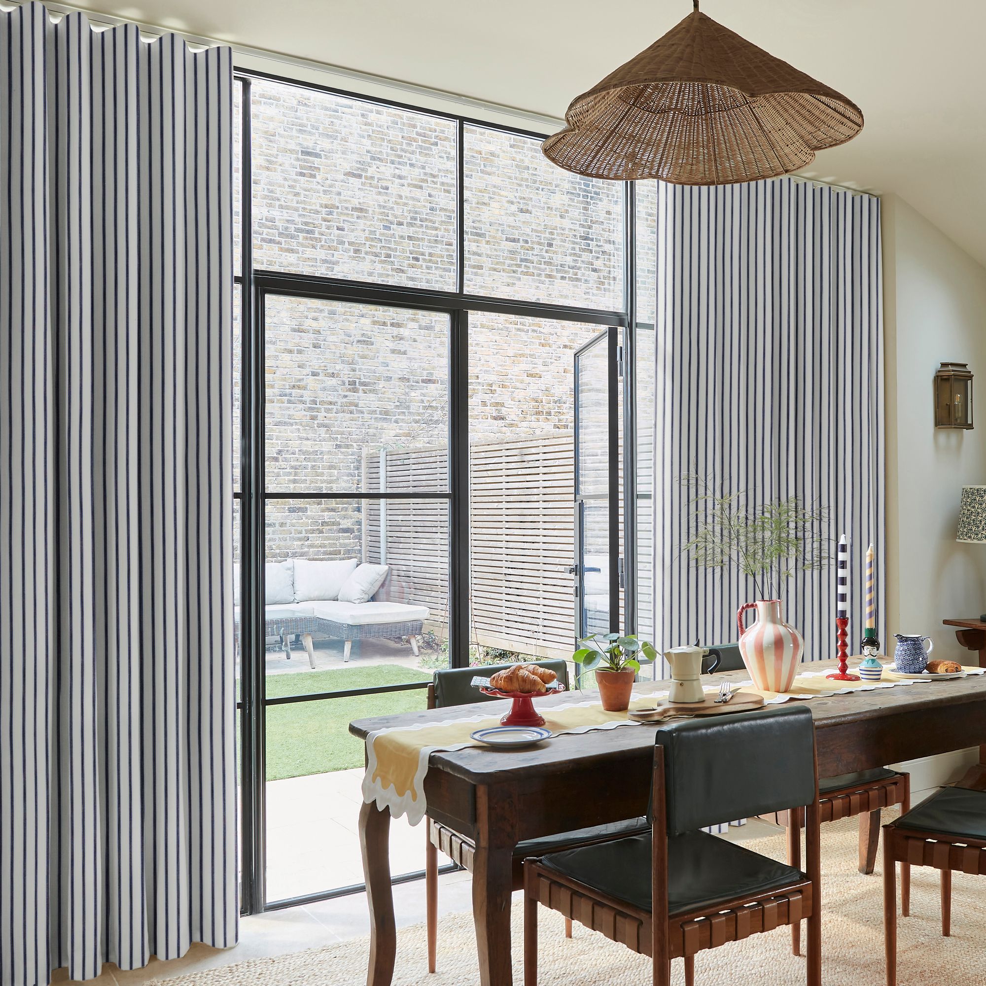 A dining table set with various items under a wicker lampshade, adjacent to large glass doors. Curtains frame the view of an outdoor sofa and brick wall courtyard.