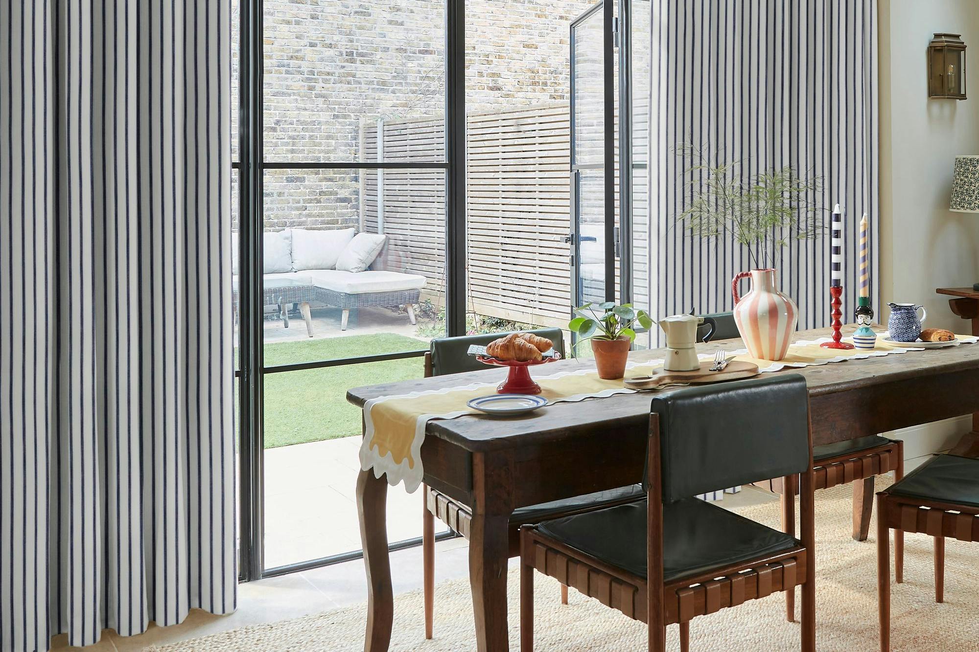 A dining table set with various items under a wicker lampshade, adjacent to large glass doors. Curtains frame the view of an outdoor sofa and brick wall courtyard.