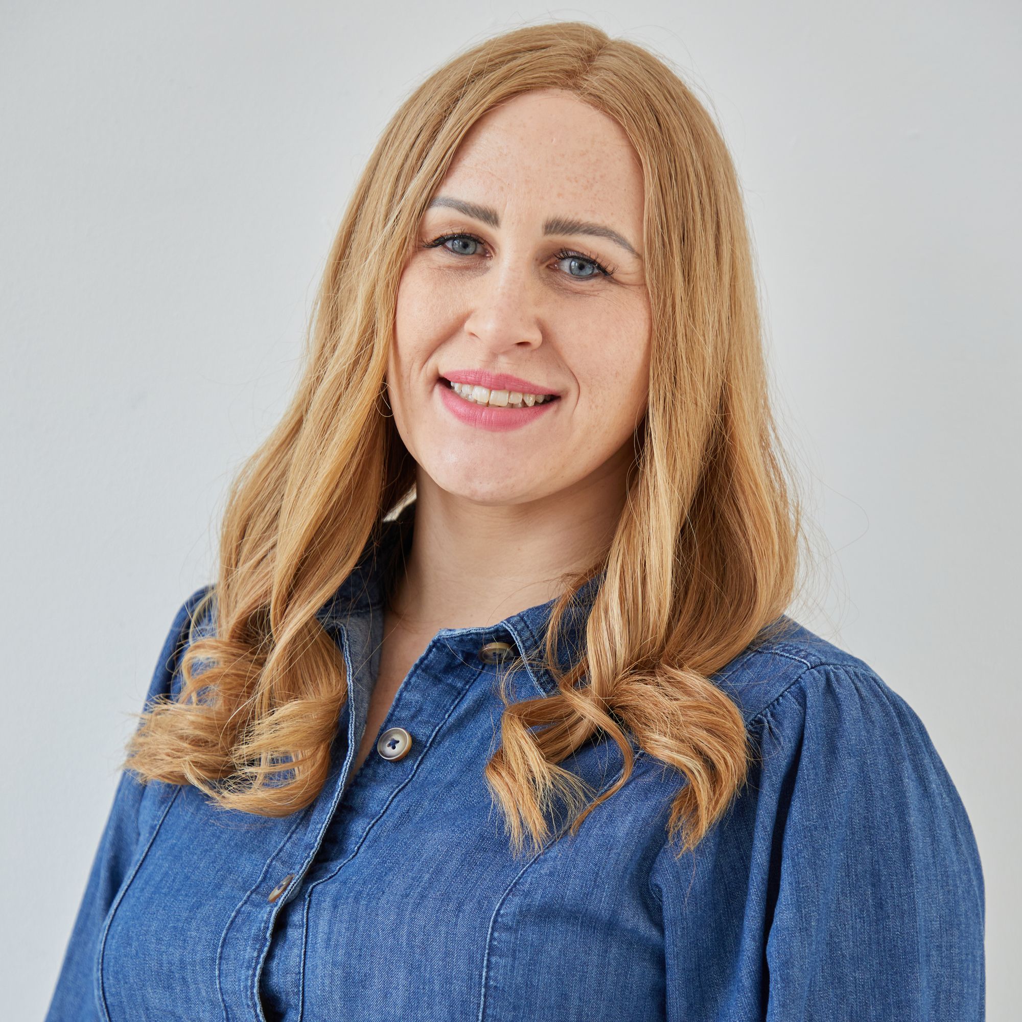 A person with long, wavy blonde hair is smiling, wearing a blue denim shirt, in front of a plain, light-colored background.