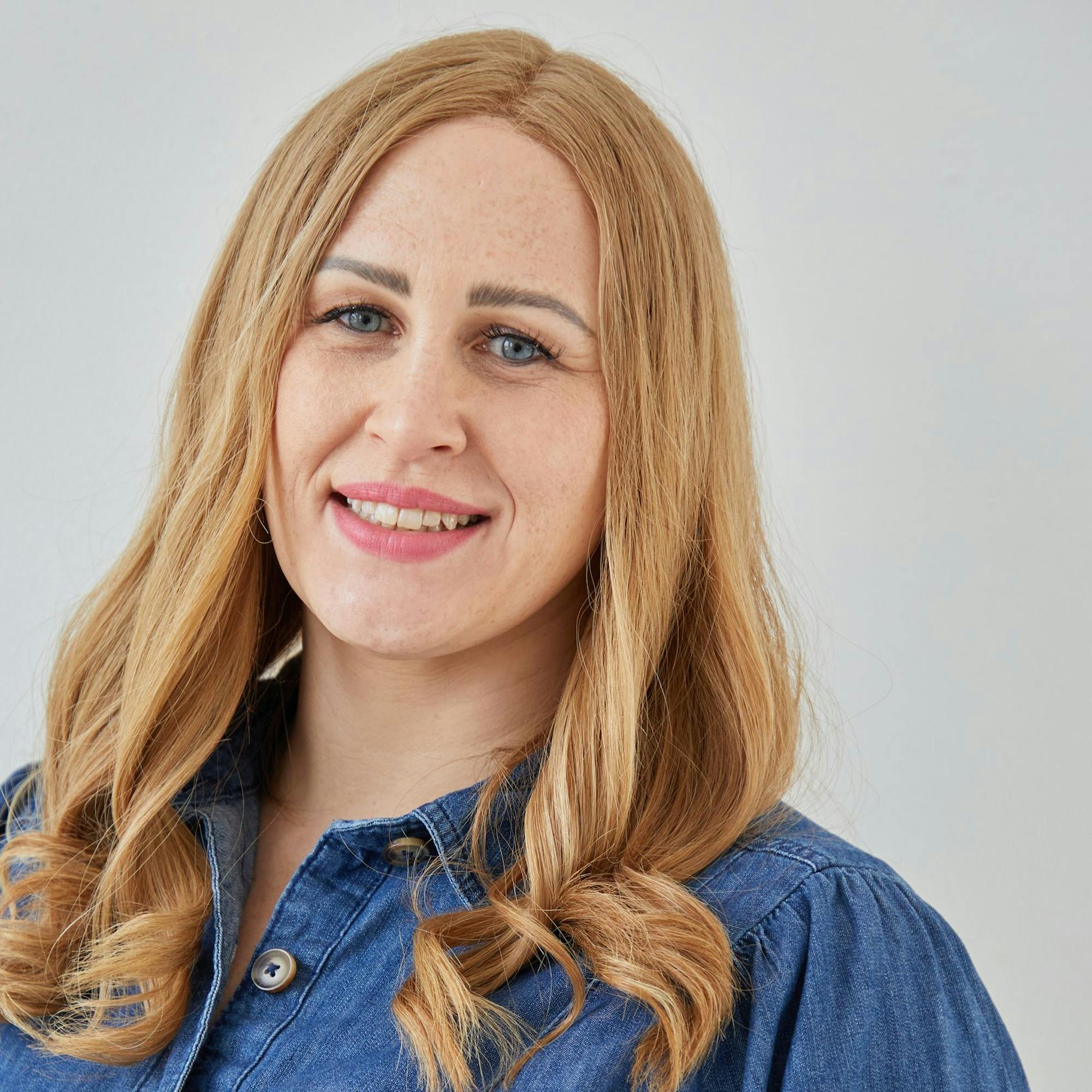 A person with long, wavy blonde hair is smiling, wearing a blue denim shirt, in front of a plain, light-colored background.