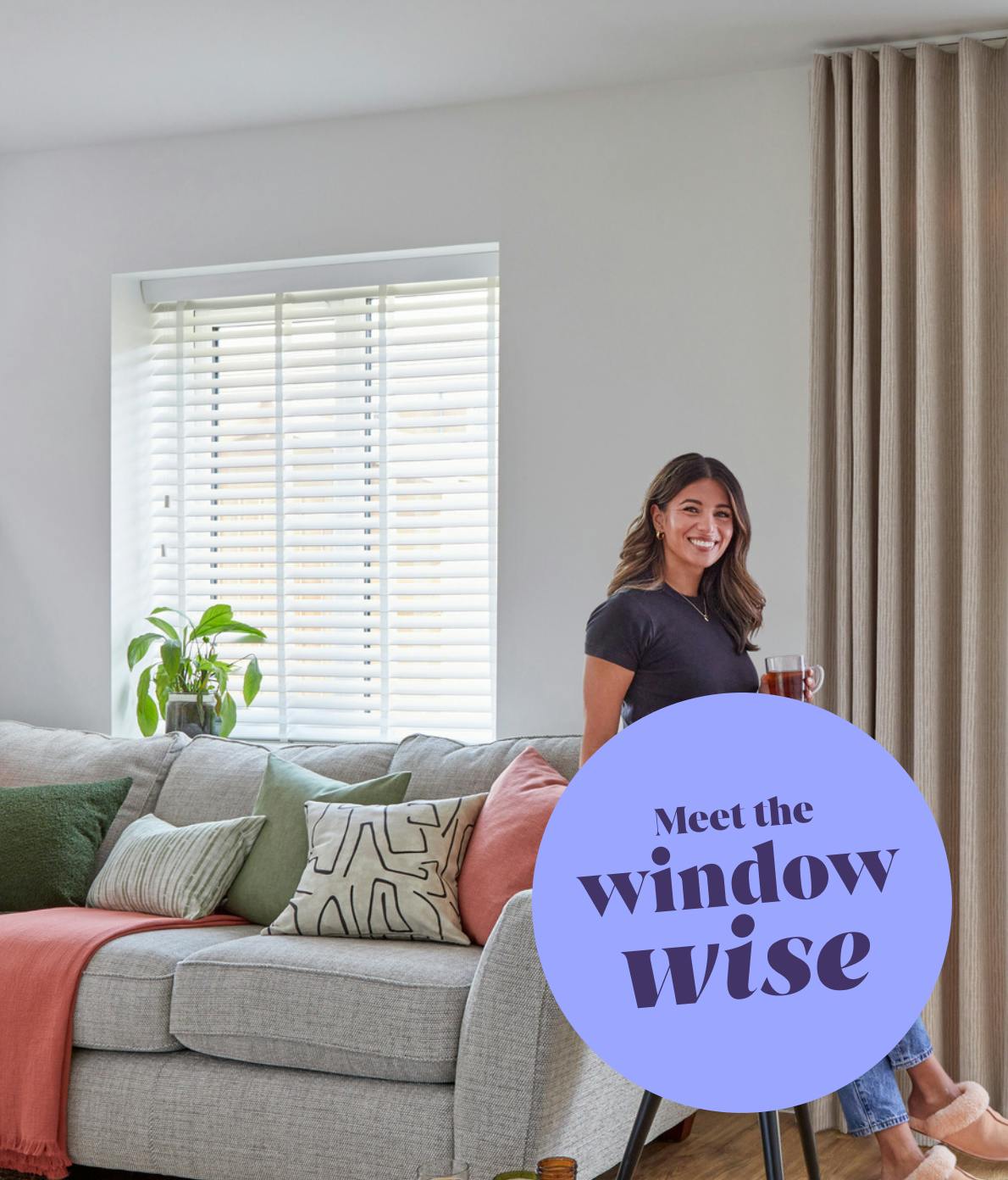 A person stands smiling, holding a mug in a living room with a gray sofa, green and peach cushions, and blinds. Large text reads "Meet the window wise."