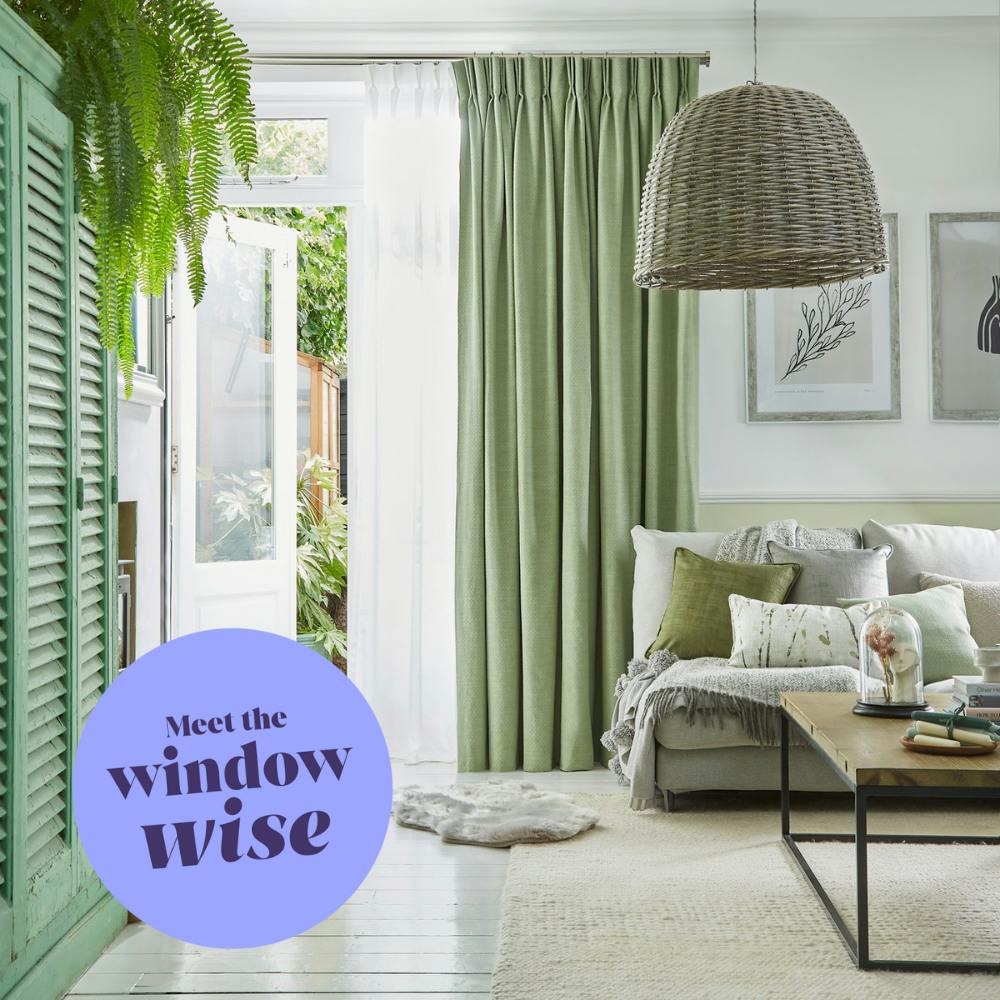 A cozy living room with a sofa, cushions, and a wooden table under a wicker lamp. Green curtains and an open door reveal a garden. Text reads "Meet the window wise."