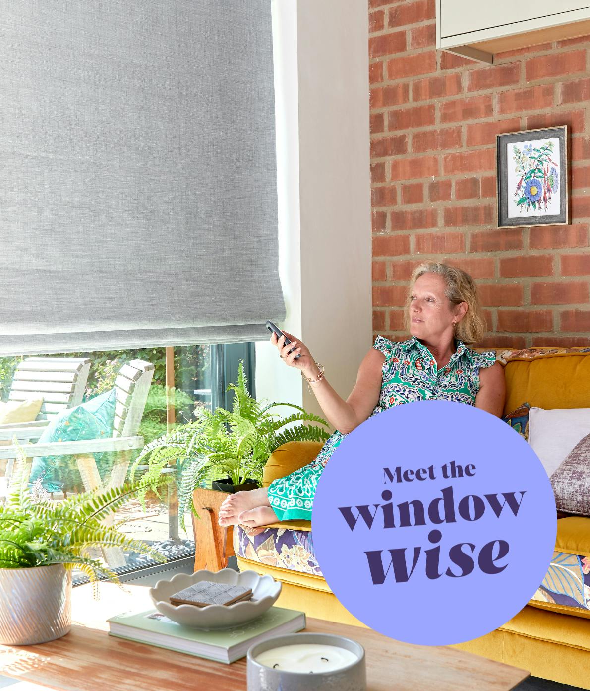 A woman relaxes on a yellow sofa, using a remote control to adjust a window blind in a sunlit living room with brick walls, plants, and outdoor view. Text: �Meet the window wise.�