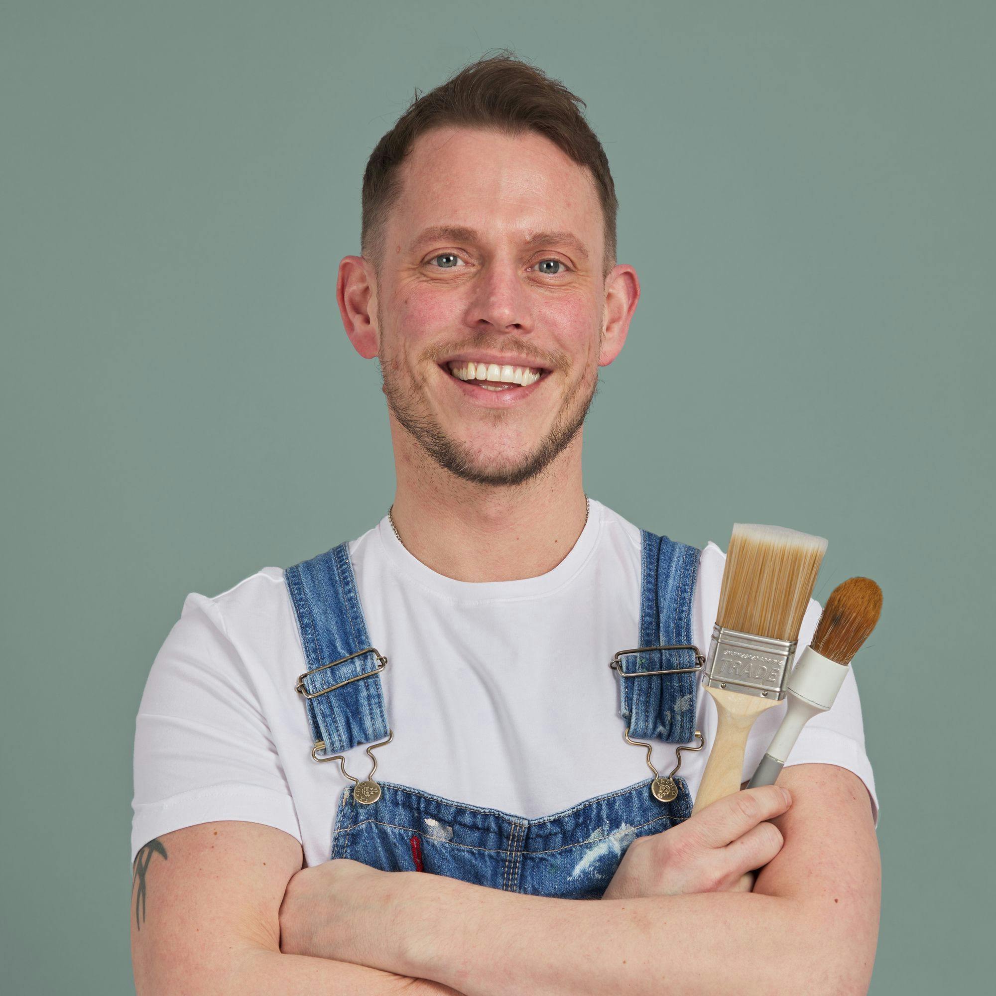 A person wearing a white T-shirt and denim overalls, holding paintbrushes while smiling confidently against a plain gray background.