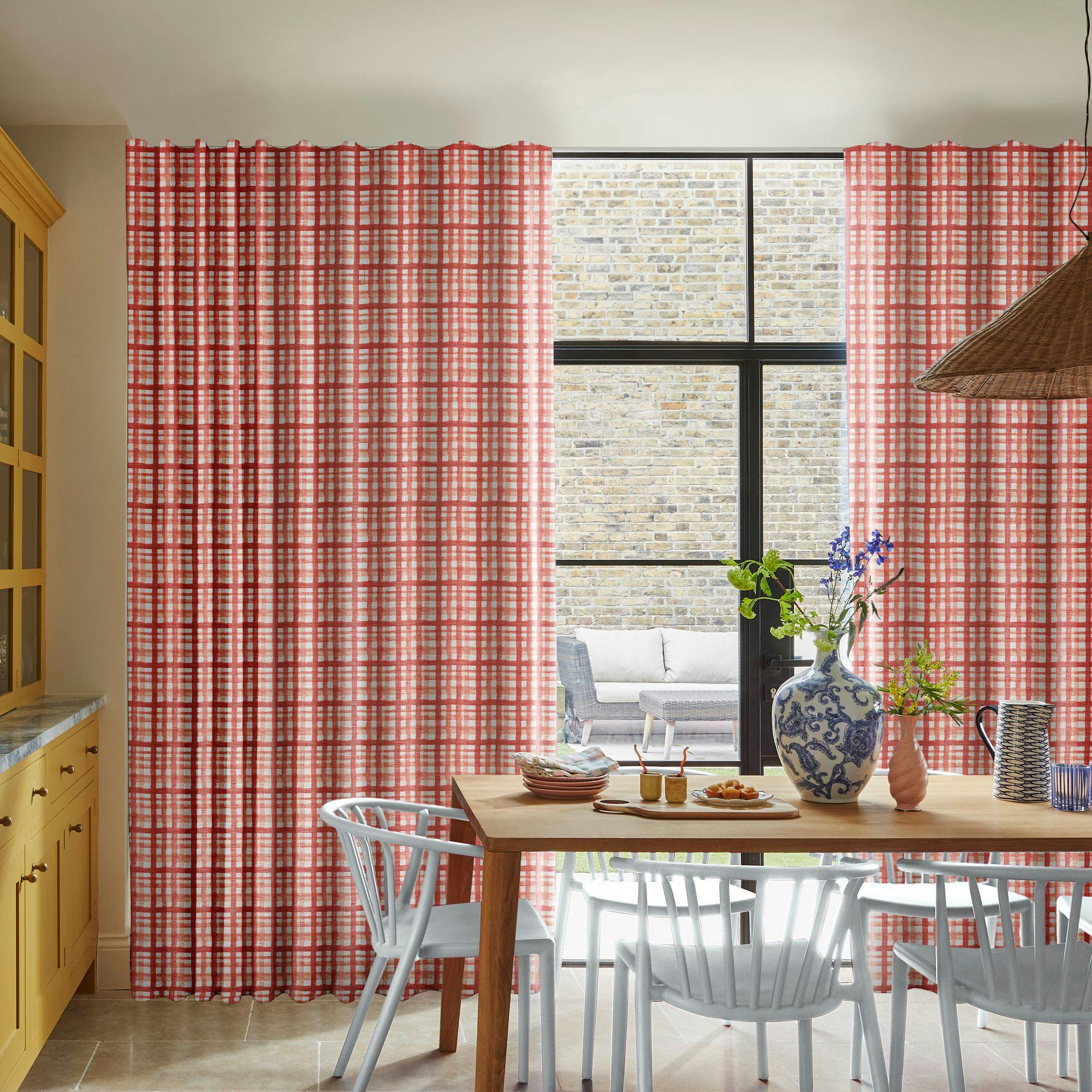Red plaid curtains hang beside a tall window, revealing a brick wall outside. A wooden dining table is set with dishes and a large blue-and-white vase, surrounded by light blue chairs.