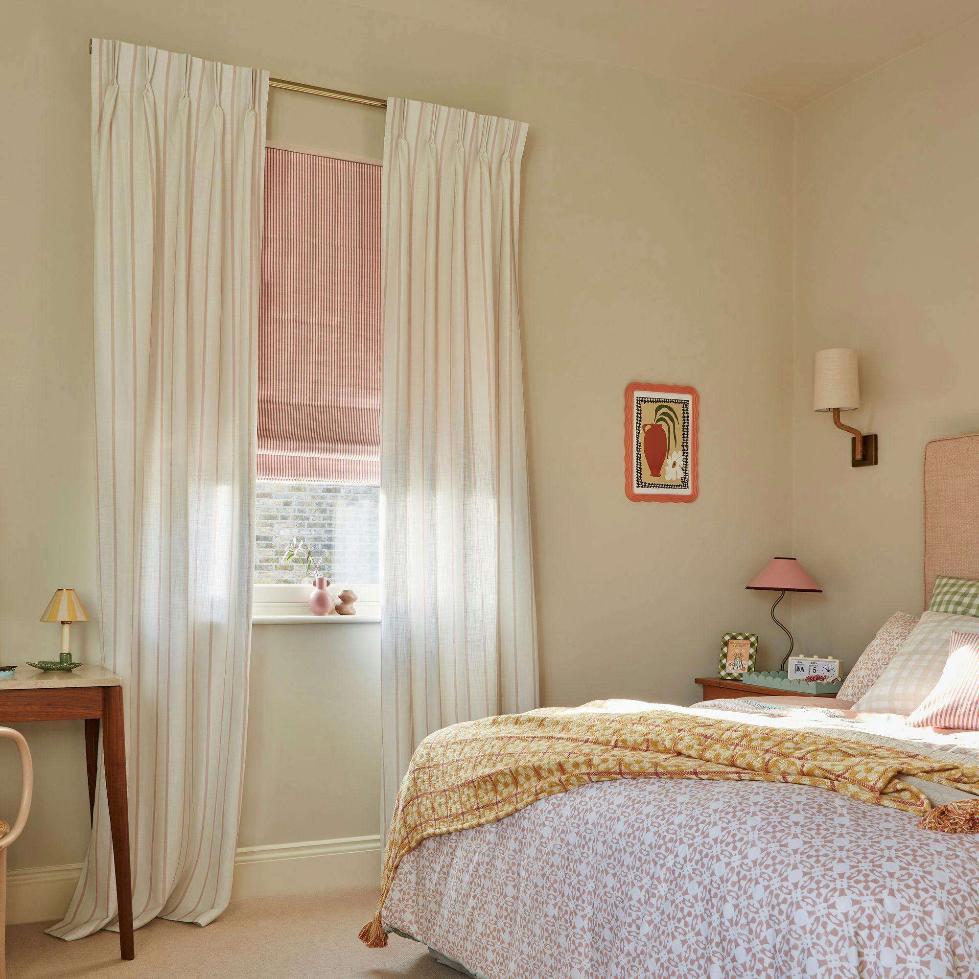 Bedroom features a neatly made bed with patterned bedding and throw, next to a nightstand with a lamp. A window with curtains and a blind allows natural light, framed by a minimalist room setting.