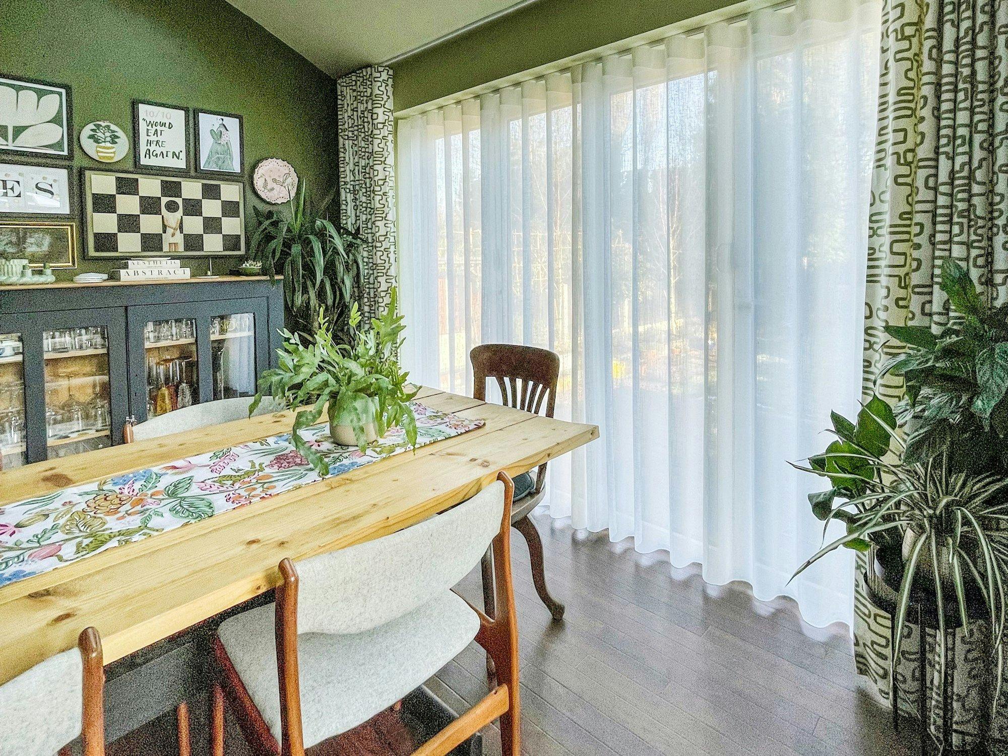 A dining table with a floral runner and a potted plant sits near large windows with sheer curtains; a dark cabinet and framed artwork adorn the green walls. Text: "10/10 would eat here again!"