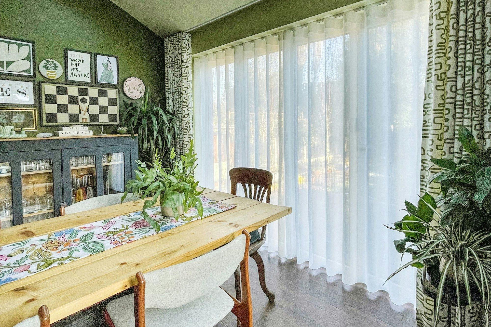 A dining table with a floral runner and a potted plant sits near large windows with sheer curtains; a dark cabinet and framed artwork adorn the green walls. Text: "10/10 would eat here again!"