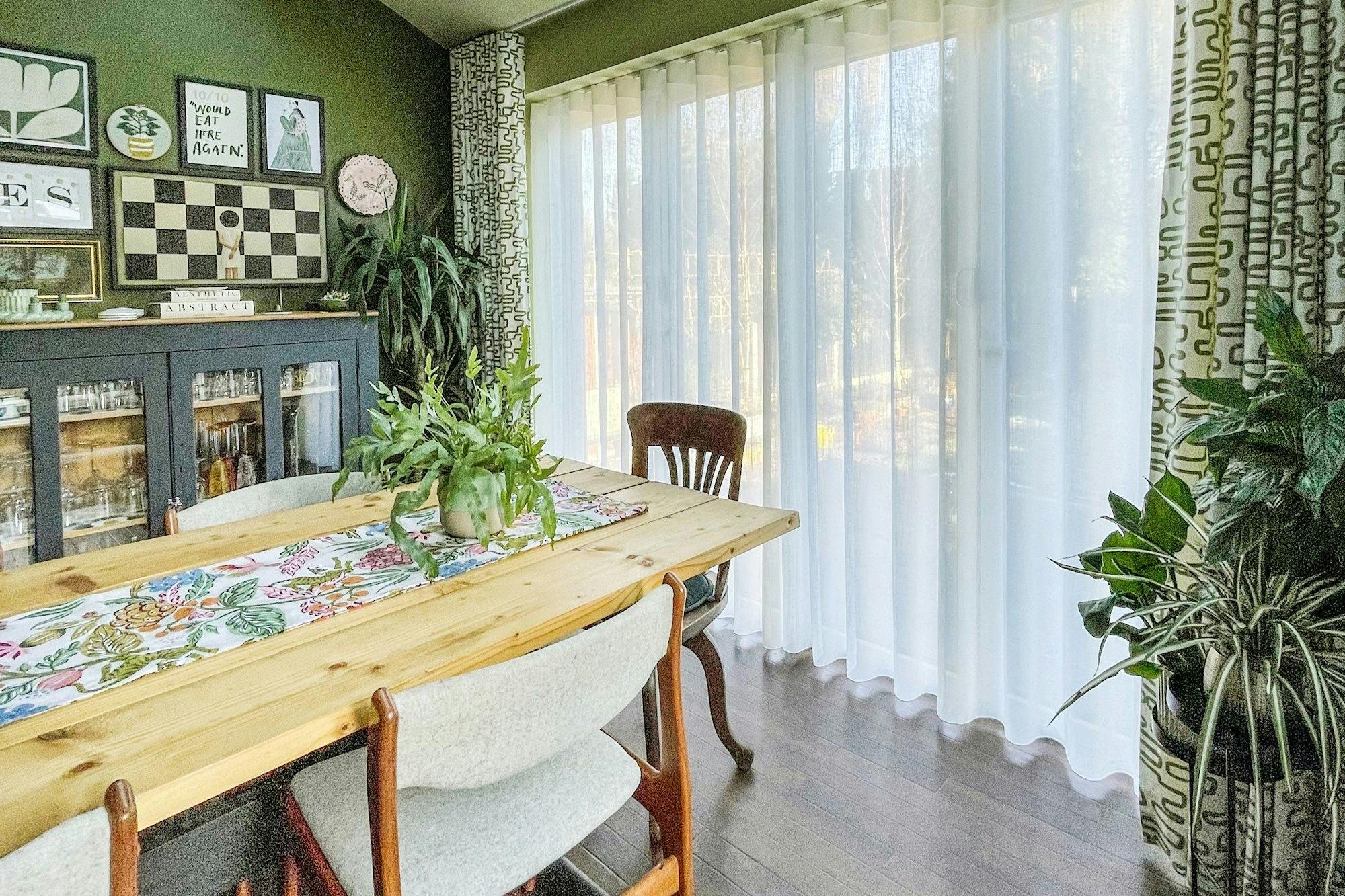 A dining table with a floral runner and a potted plant sits near large windows with sheer curtains; a dark cabinet and framed artwork adorn the green walls. Text: "10/10 would eat here again!"