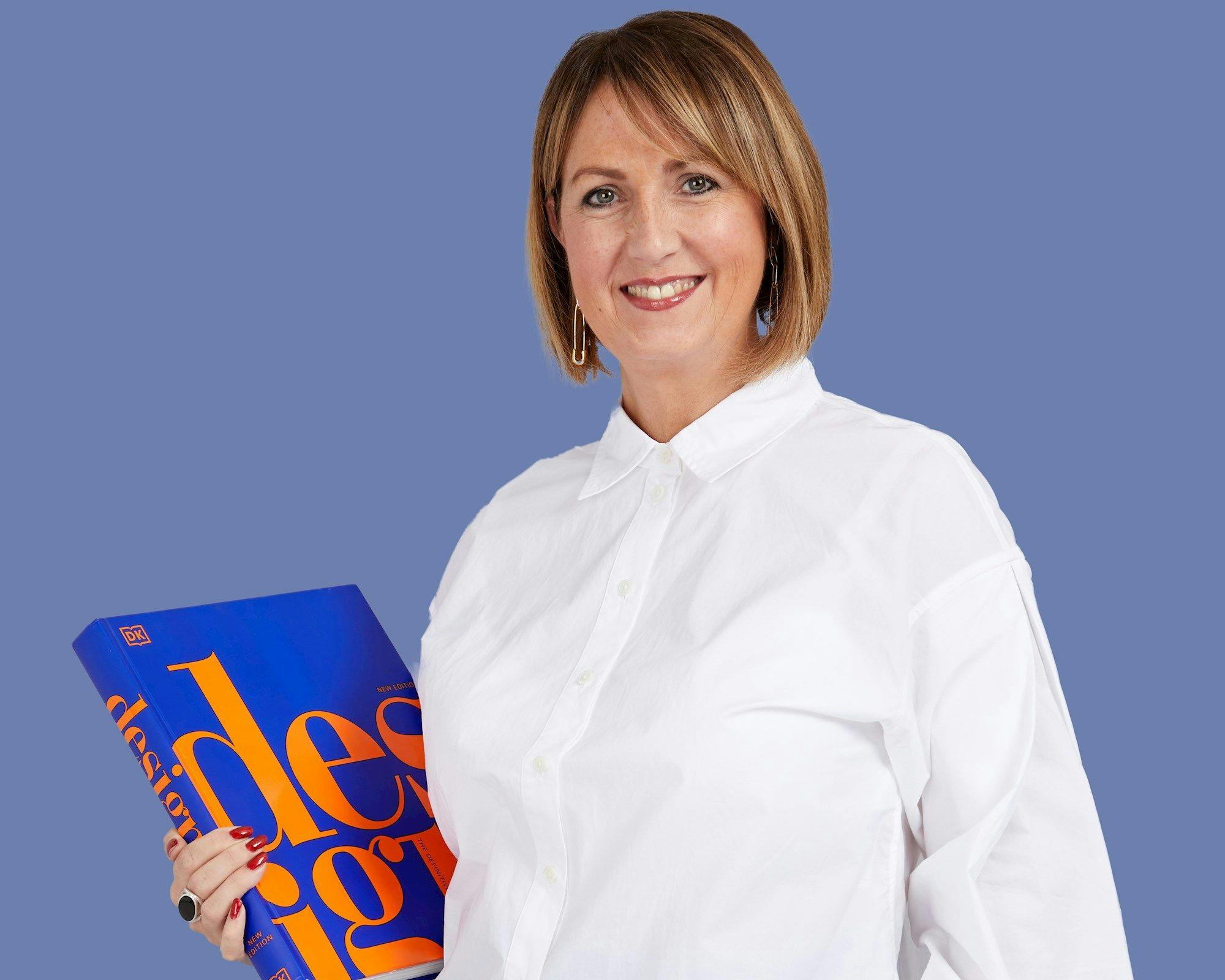 A person wearing a white shirt holds a large blue book titled "design" in orange letters, set against a plain blue background. They are smiling and wearing hoop earrings.