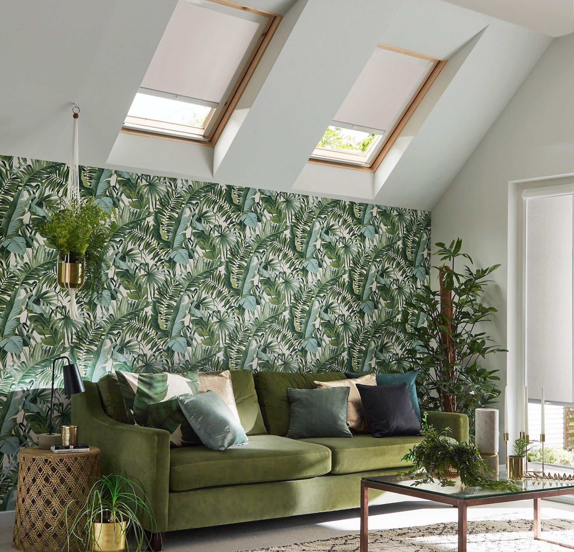 A green sofa adorned with multicolored cushions sits against a tropical leaf-patterned wallpaper in a sunlit room with skylights. Surrounding the sofa are plants and a glass coffee table.