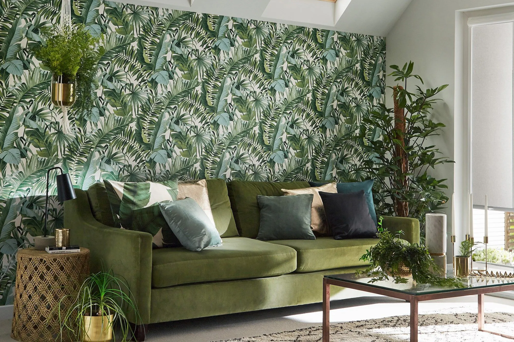 A green sofa adorned with multicolored cushions sits against a tropical leaf-patterned wallpaper in a sunlit room with skylights. Surrounding the sofa are plants and a glass coffee table.