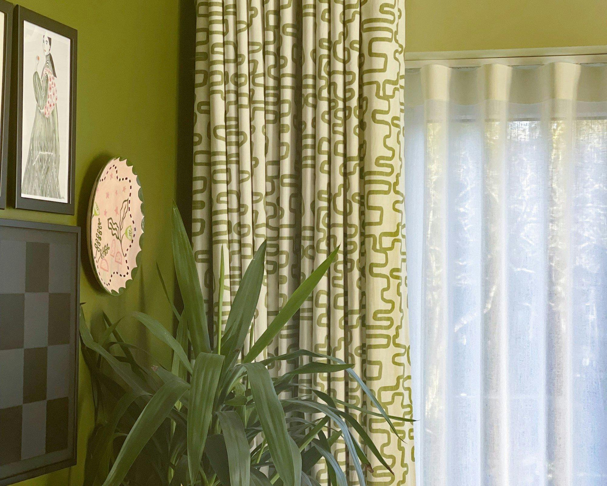 A potted plant with long, green leaves stands beside patterned curtains; wall adorned with an abstract checkered painting, a framed figure drawing, and a decorative pink plate.