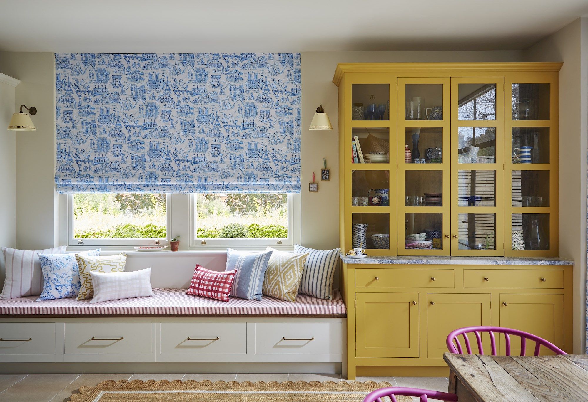 Window seat with striped bench and patterned pillows; action: situated beneath a decorative blue-patterned blind; context: adjacent to a yellow cabinet displaying dishes, in a brightly lit room.