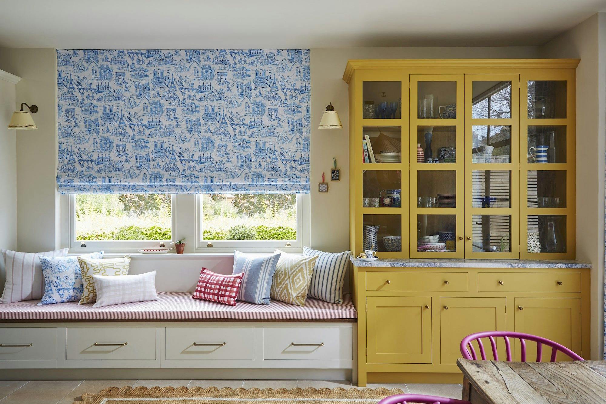 Window seat with striped bench and patterned pillows; action: situated beneath a decorative blue-patterned blind; context: adjacent to a yellow cabinet displaying dishes, in a brightly lit room.