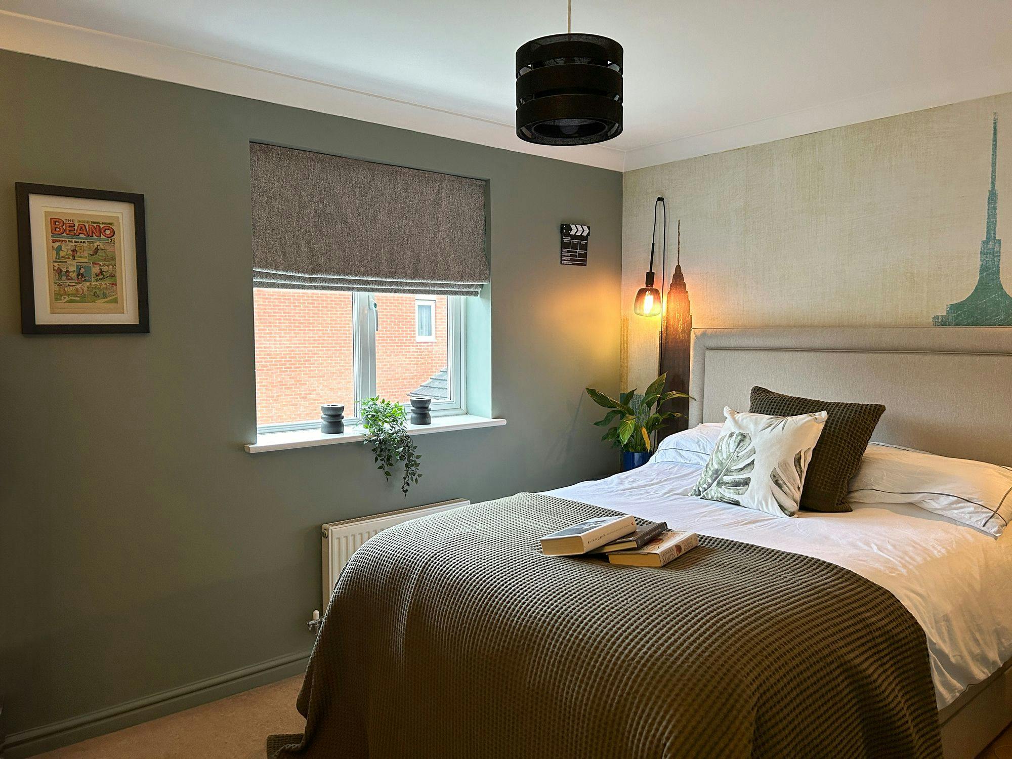 A neatly made bed with green and white pillows, adorned with books, sits beneath a pendant light in a cozy bedroom. The walls feature framed art and a window with plants. Text: "BEANO".