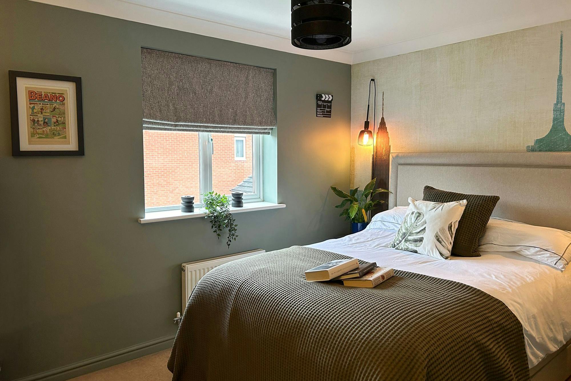 A neatly made bed with green and white pillows, adorned with books, sits beneath a pendant light in a cozy bedroom. The walls feature framed art and a window with plants. Text: "BEANO".