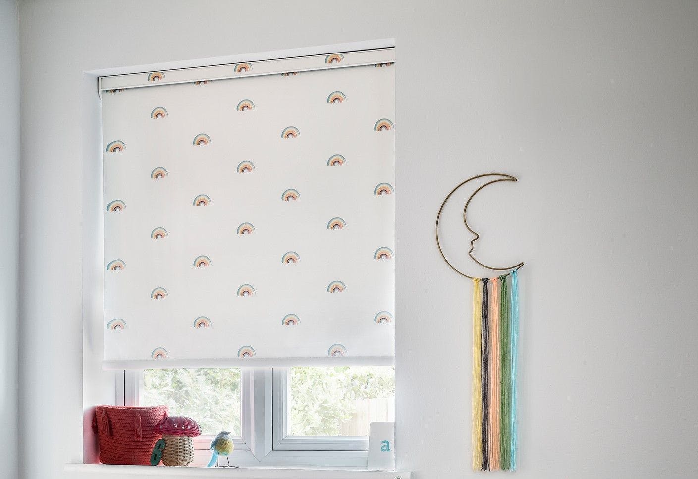 A window features a rainbow-patterned blind, partially raised, surrounded by woven baskets. Nearby, a decorative crescent moon hangs with colorful yarn strands cascading down.