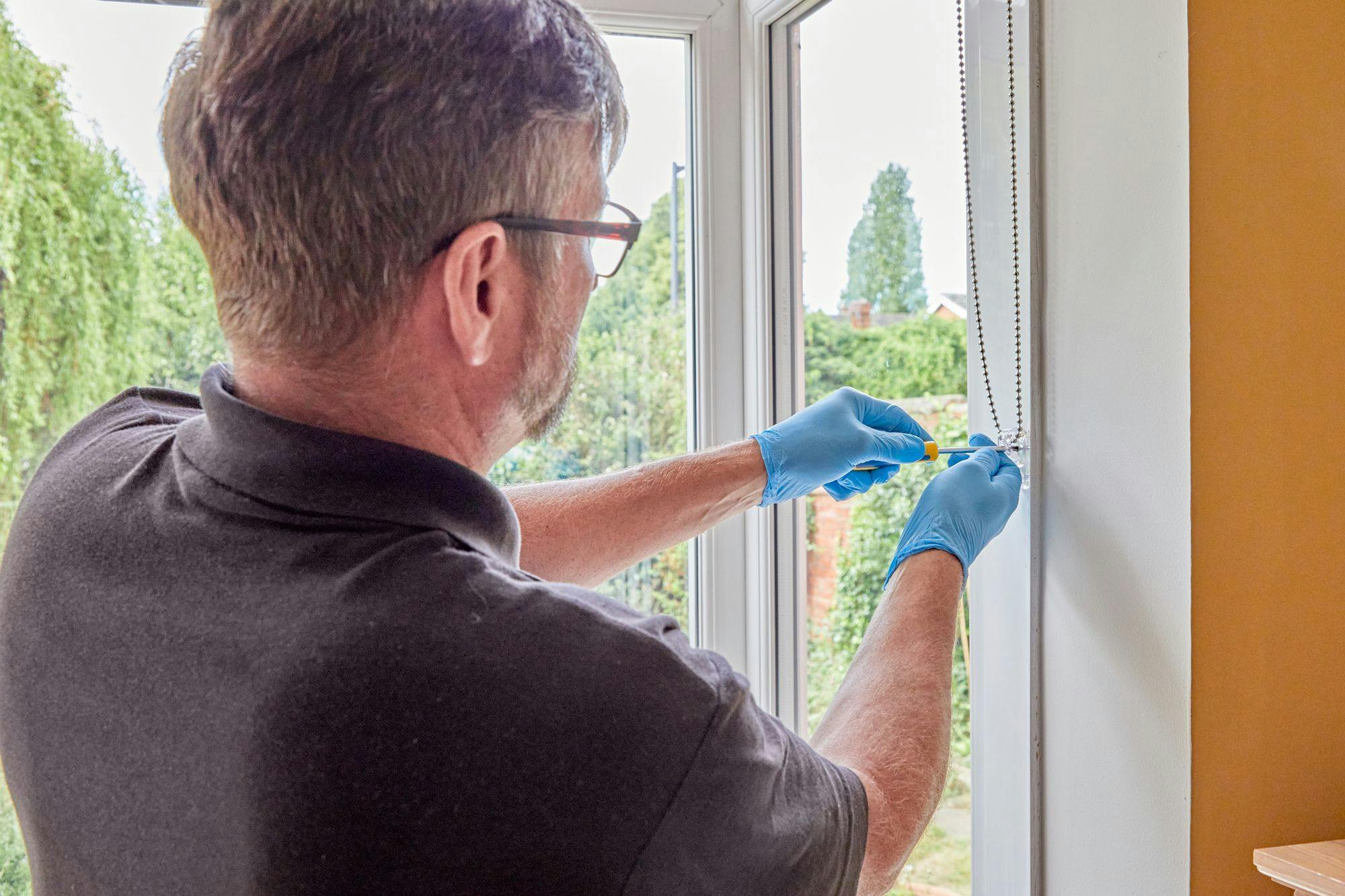 A person in a black shirt and blue gloves screws a fixture to a window frame. The action occurs indoors, with a view of lush greenery outside the window.