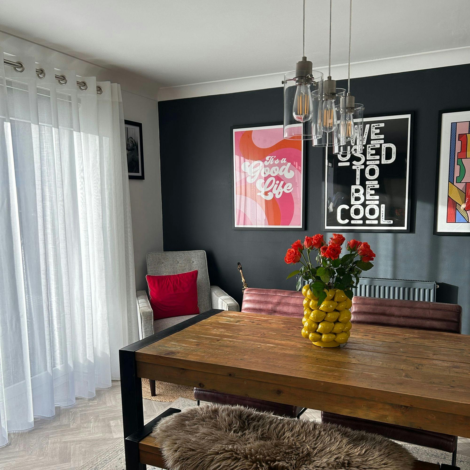 A wooden table holds a vase of red roses, while nearby a gray chair with a red cushion sits under wall art saying �It�s a Good Life� and �We Used to Be Cool.�