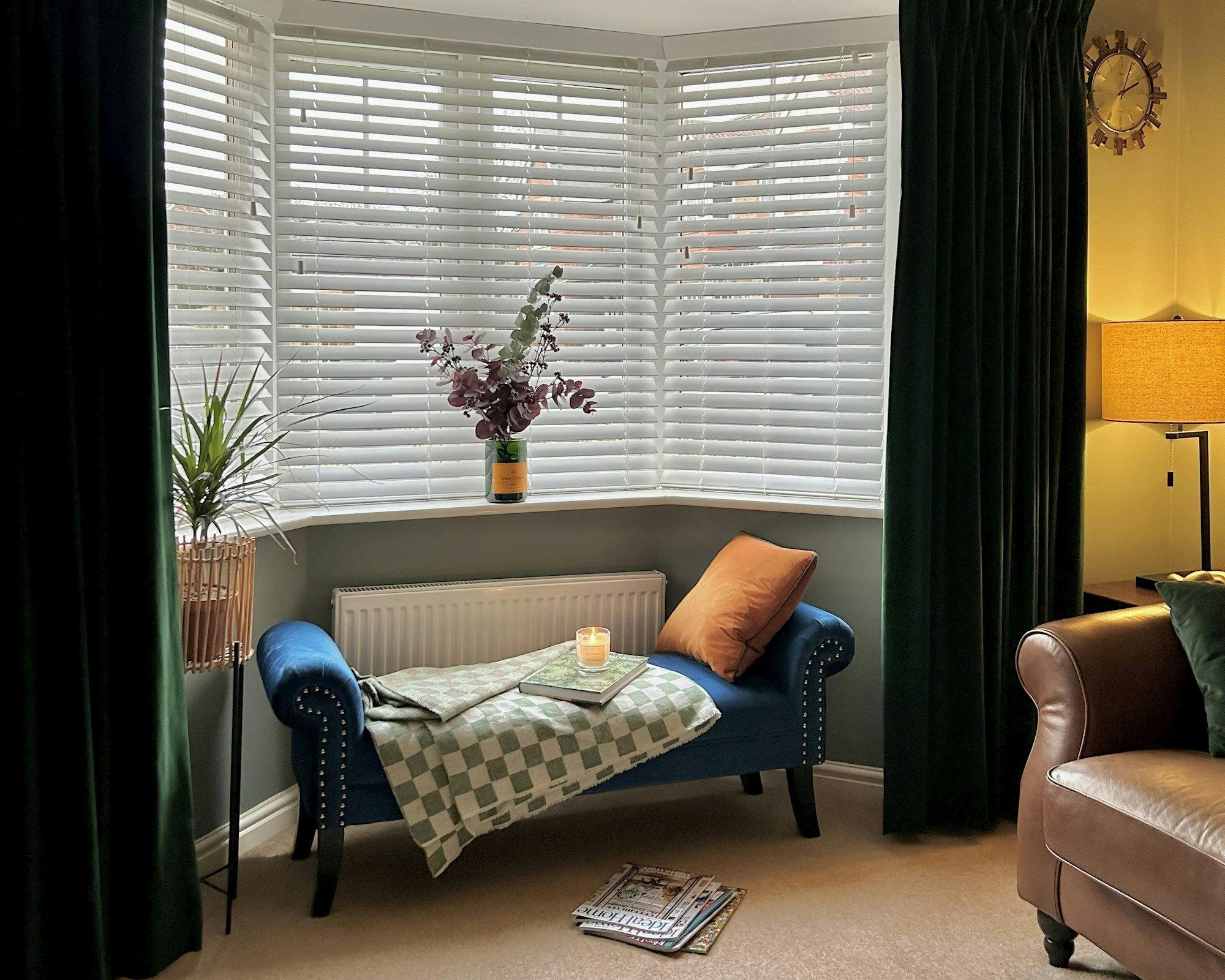 A cozy living room features a blue chaise lounge with pillows and a blanket, set near a window with blinds; a plant and candle create a relaxed atmosphere.