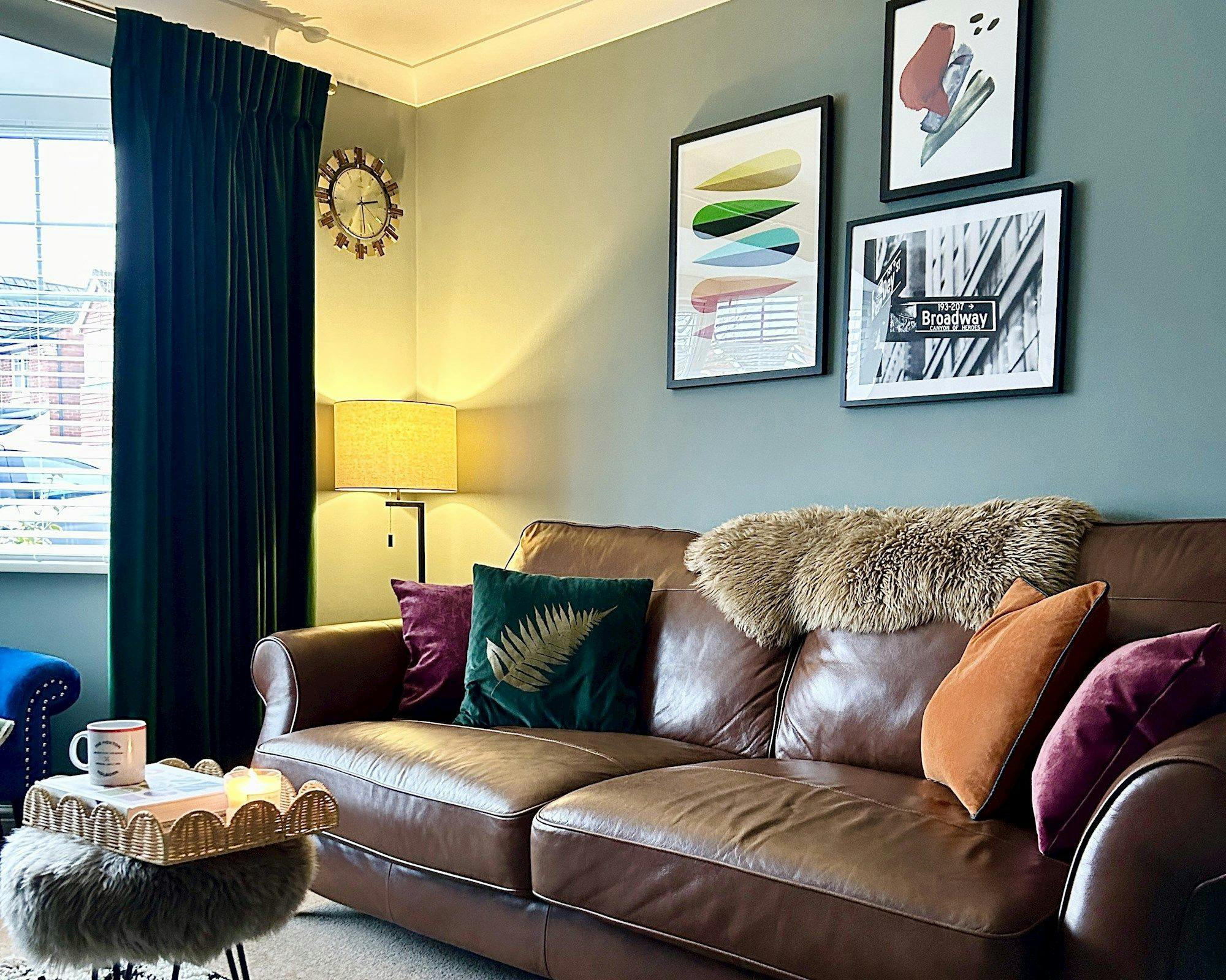 A brown leather sofa adorned with colorful cushions and a fur throw sits against a wall with framed artwork, including a "Broadway" sign. Nearby, a lit lamp and clock enhance the cozy living room.