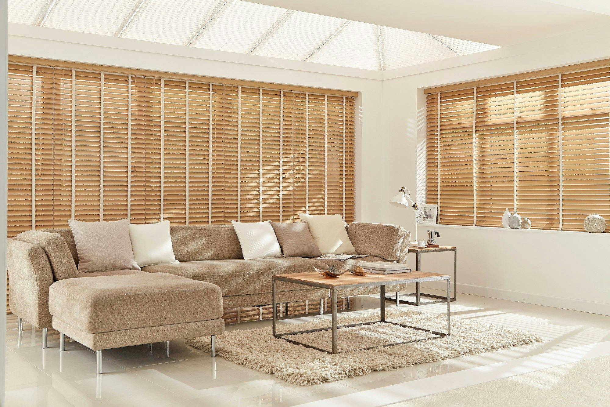 A beige sectional sofa sits with white pillows in a bright room. Wooden blinds cover windows, and a coffee table with decor rests on a textured rug, surrounded by minimal decor.