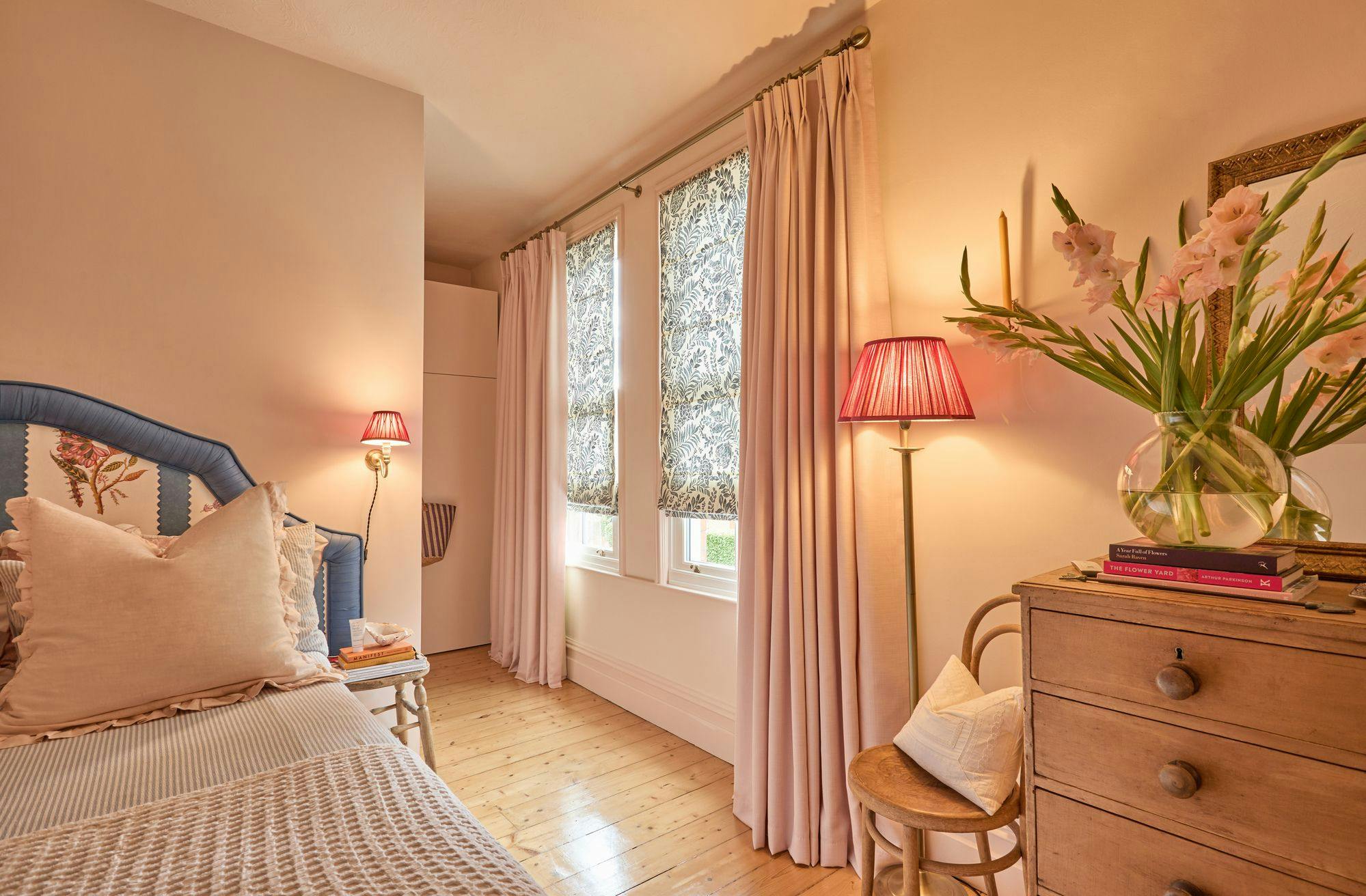 Bed with floral headboard and cushions rests beside a bedside table with books. Pink lamps and floral curtains enhance the wooden-floored room's cozy ambiance. Vase with flowers atop a dresser.