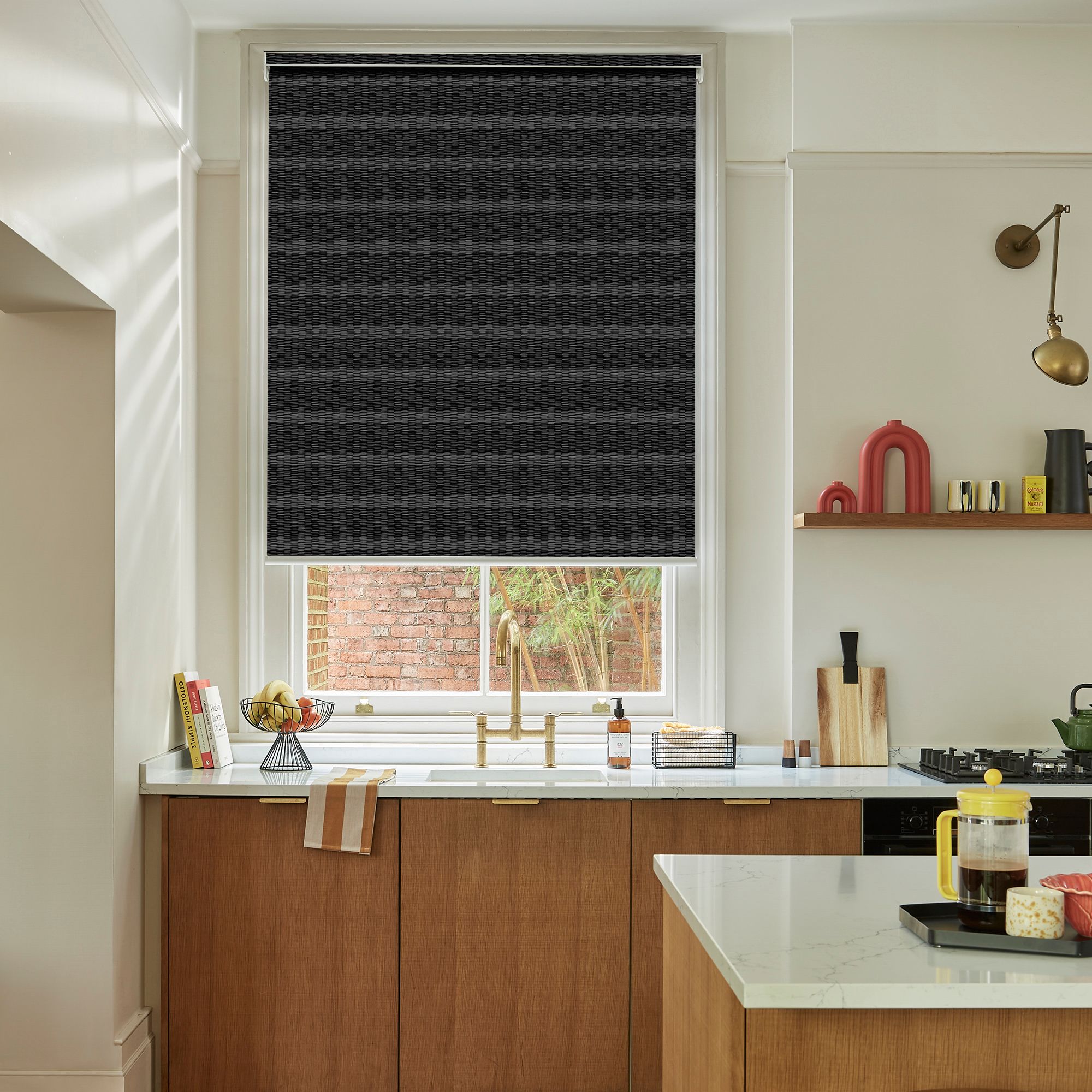 A kitchen features a black roller blind covering a window above a wooden sink area. Countertops and shelves hold assorted kitchen items like books, bowls, utensils, and a yellow French press.