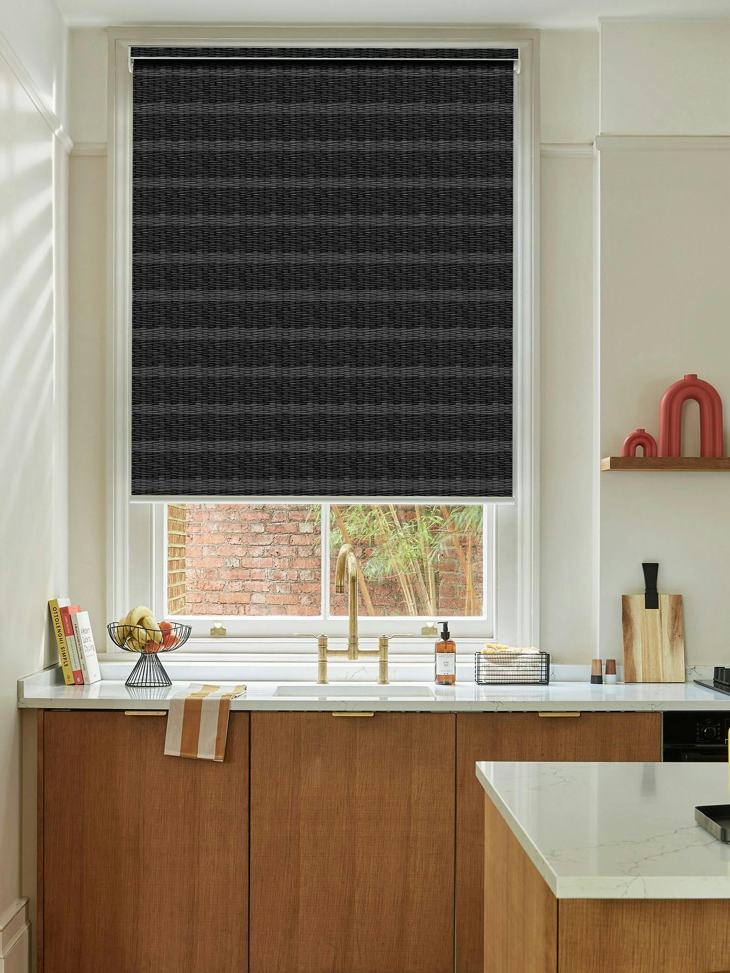 A kitchen features a black roller blind covering a window above a wooden sink area. Countertops and shelves hold assorted kitchen items like books, bowls, utensils, and a yellow French press.