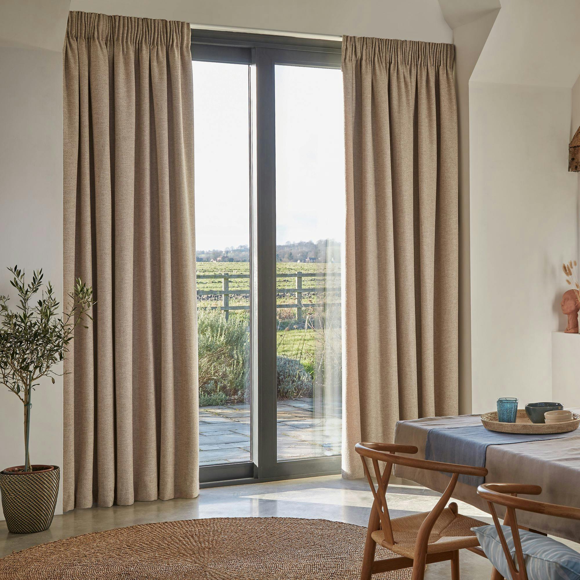Beige curtains hang beside a large sliding glass door, revealing a view of a green field and wooden fence. A dining table with chairs and a woven rug complete the cozy interior setting.