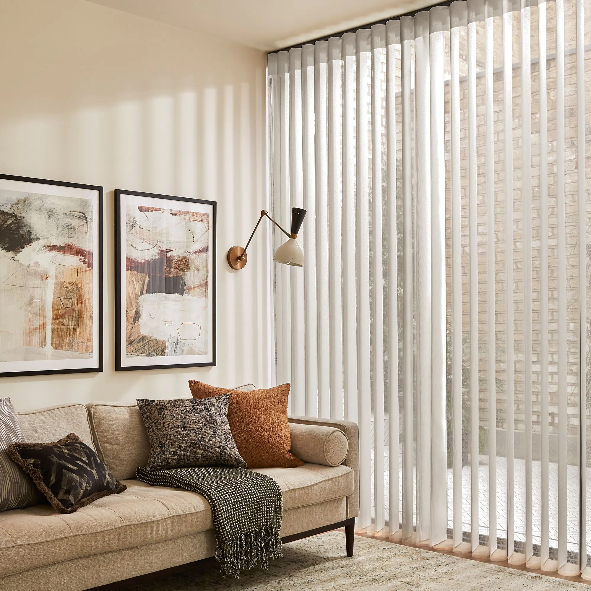 A beige couch with patterned cushions and a throw sits below two abstract paintings. A modern wall lamp illuminates a room with floor-to-ceiling sheer curtains, softly filtering daylight.