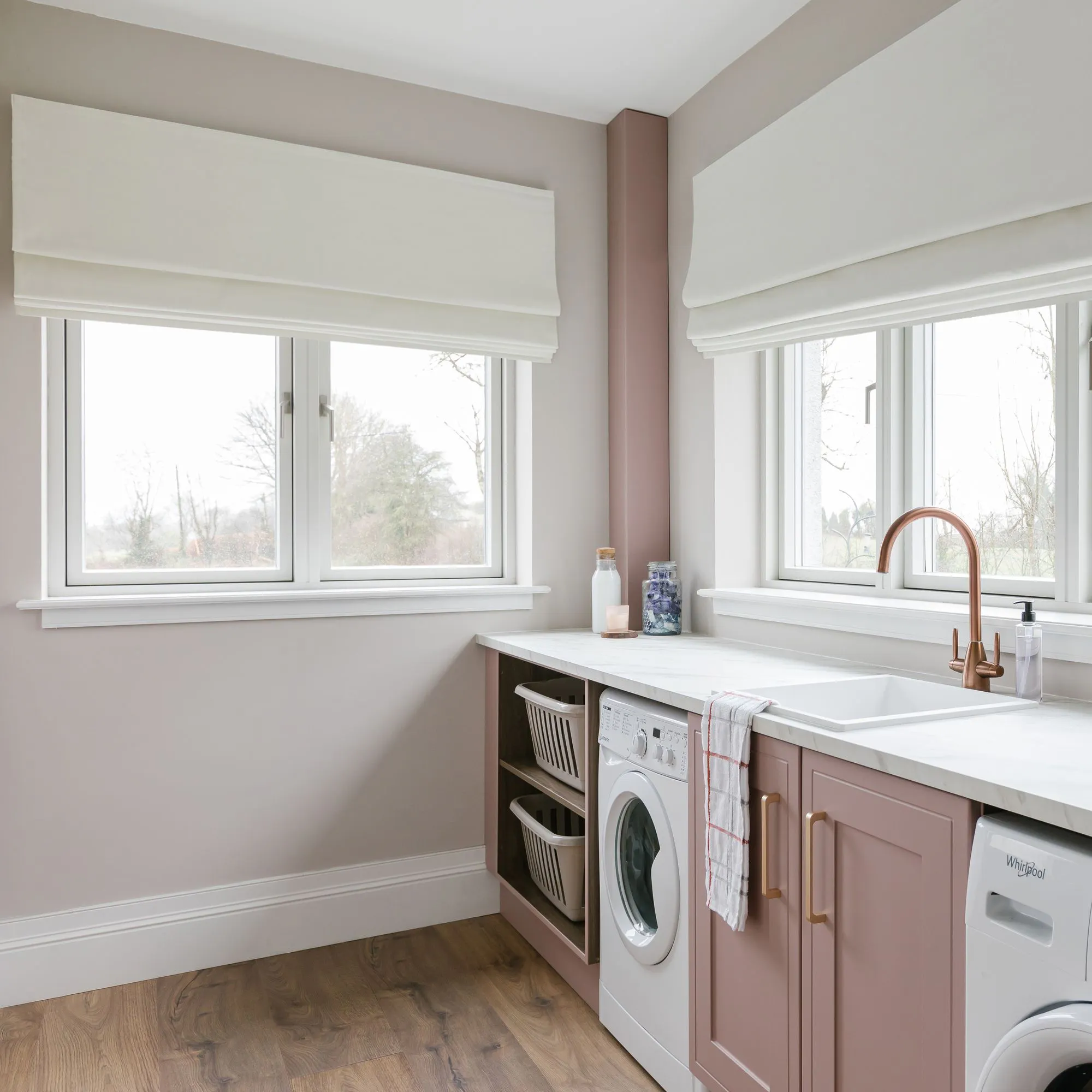 A laundry room features a washing machine beneath a white countertop with pink cabinets. Large windows with beige roman shades provide natural light. The wooden floor adds warmth to the space.