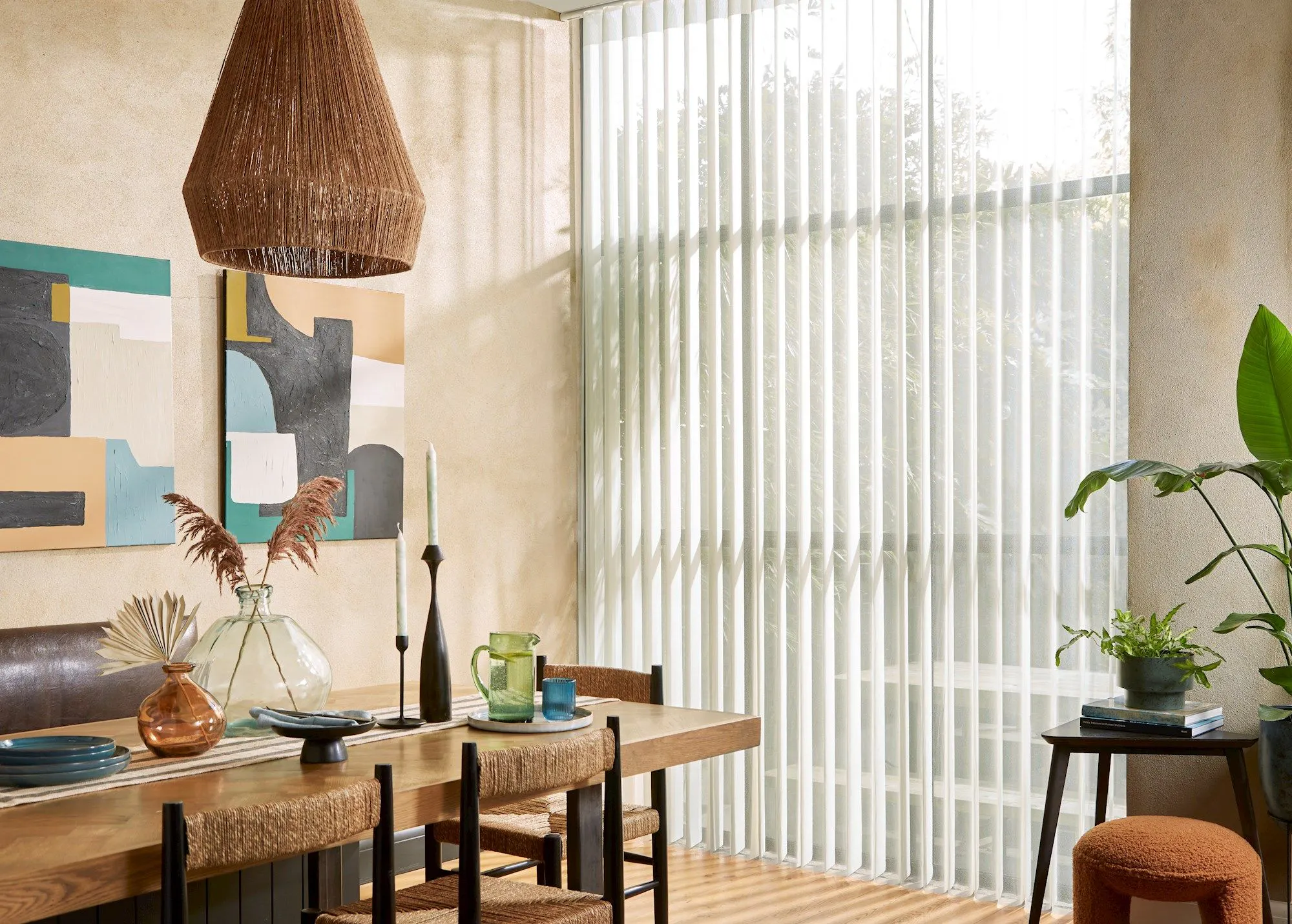 Dining room with floor to ceiling windows and doors dressed in pale grey diffused sheer full-length blinds, tilted wooden table with raffia chairs, raffia shade on pendant light, abstract art in shades of blue and brown on the brown marble-effect walls.