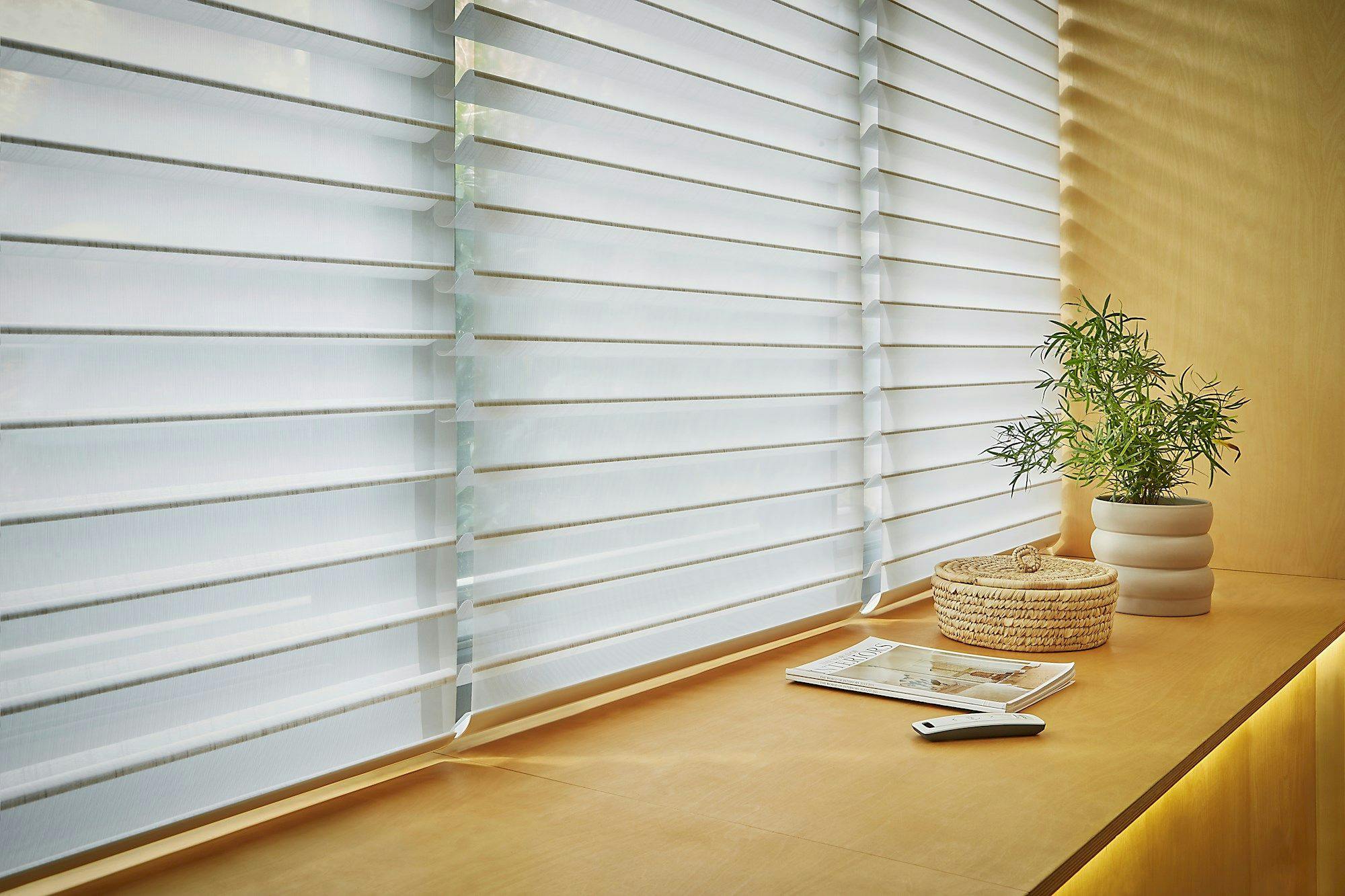 Close up of three pale grey triple shade sheer blinds, fully lowered and vanes slightly tilted in a deep window recess in pale terracotta, slimline white remote control, interior magazing, small spiny plant in a pale beige ribbed pot and a small raffia basket all on the sill.