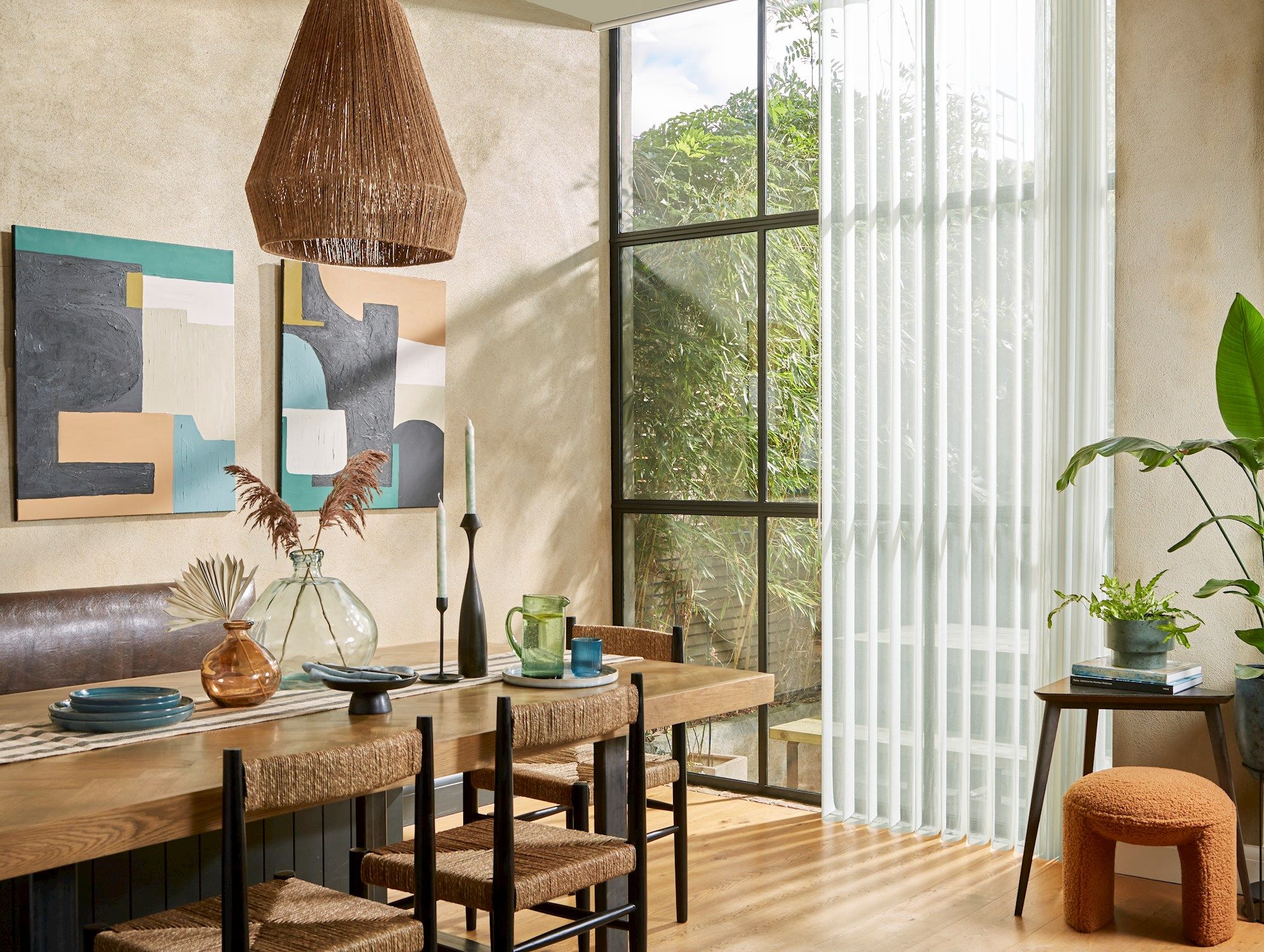 A dining table with woven chairs holds decorative vases and tableware, beneath a basket-like pendant light. Abstract art and large windows with sheer curtains enhance the rustic, light-filled room.
