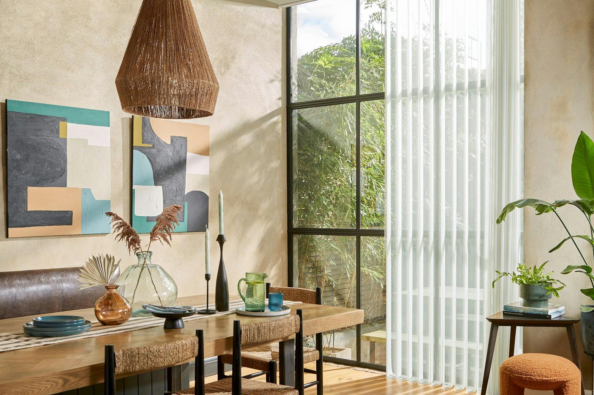A dining table with woven chairs holds decorative vases and tableware, beneath a basket-like pendant light. Abstract art and large windows with sheer curtains enhance the rustic, light-filled room.