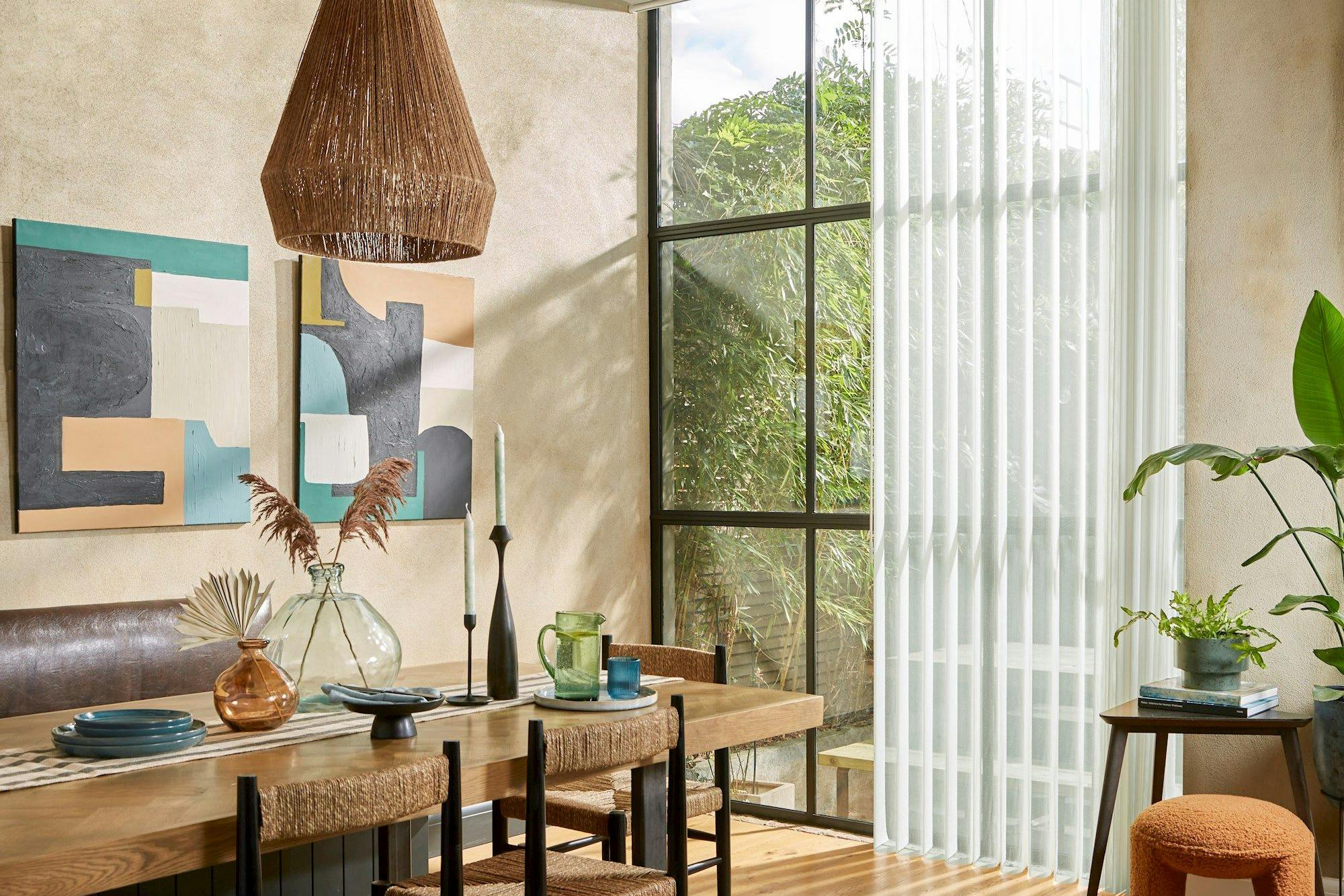 A dining table with woven chairs holds decorative vases and tableware, beneath a basket-like pendant light. Abstract art and large windows with sheer curtains enhance the rustic, light-filled room.