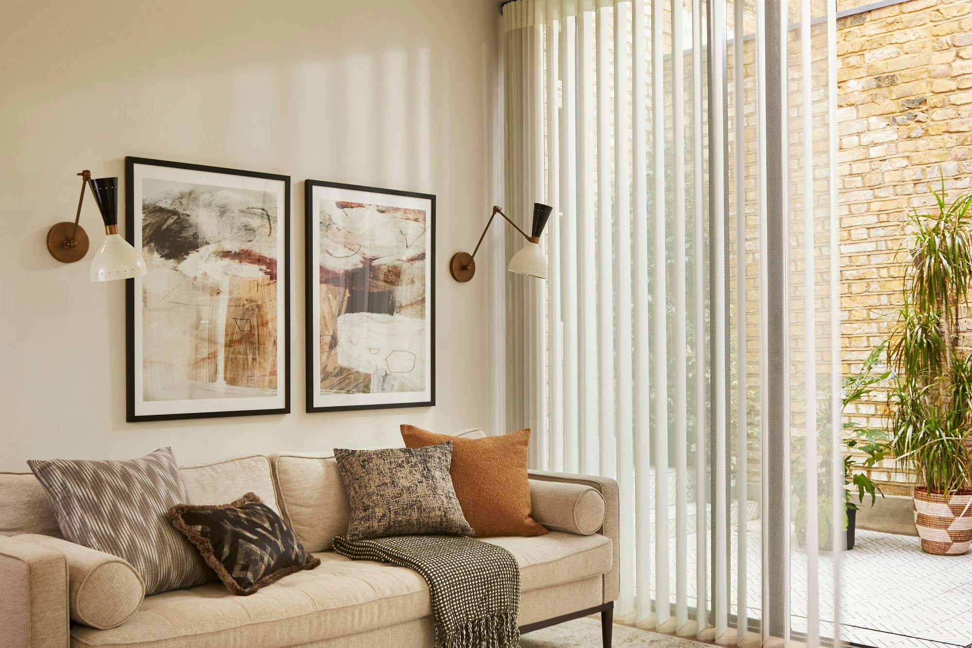 Beige couch adorned with patterned pillows and a throw blanket sits beneath abstract wall art. Adjacent, vertical blinds filter light from a brick-walled patio with potted plants.