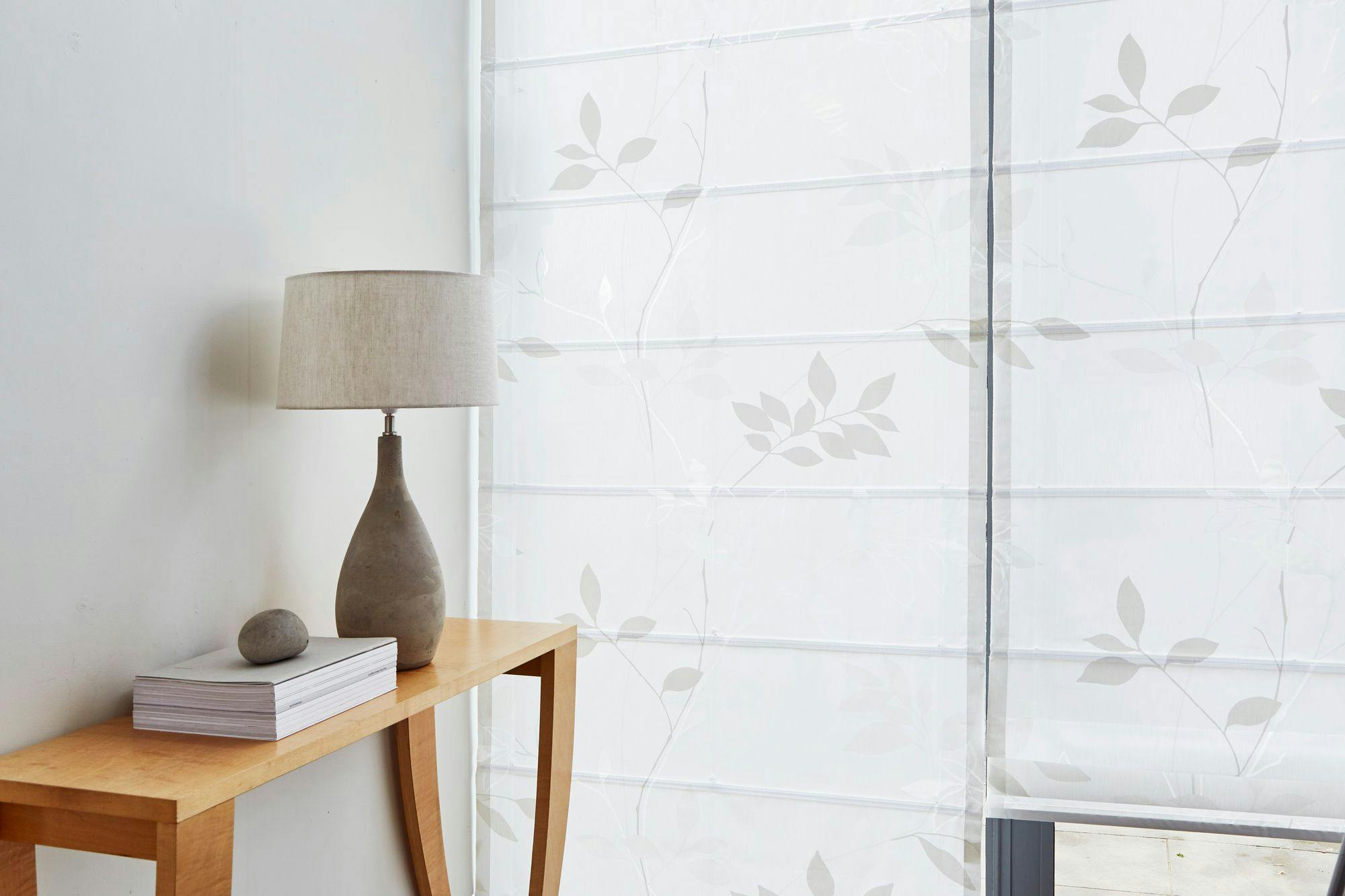 A beige table lamp sits on a wooden console alongside a stacked book and a stone. Background includes patterned sheer curtains with leaf designs filtering light.