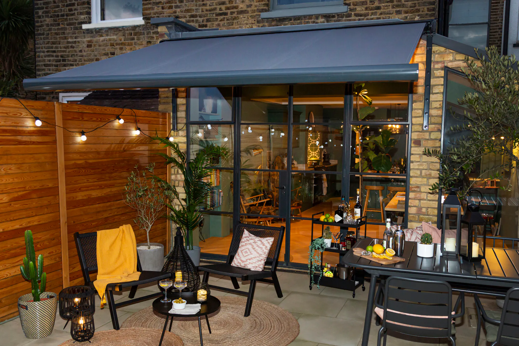 exterior small patio at dusk with black dining table and chairs to right, on left two black chairs and low table on round raffia mats, lit candles.