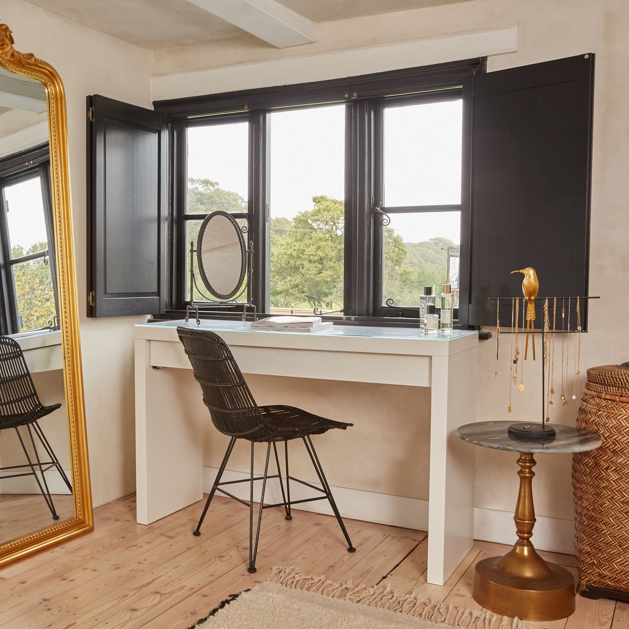 A rattan chair is positioned by a white vanity table with cosmetics, near black-framed windows overlooking greenery. A gold jewelry holder on a small table complements the cozy, elegant room.