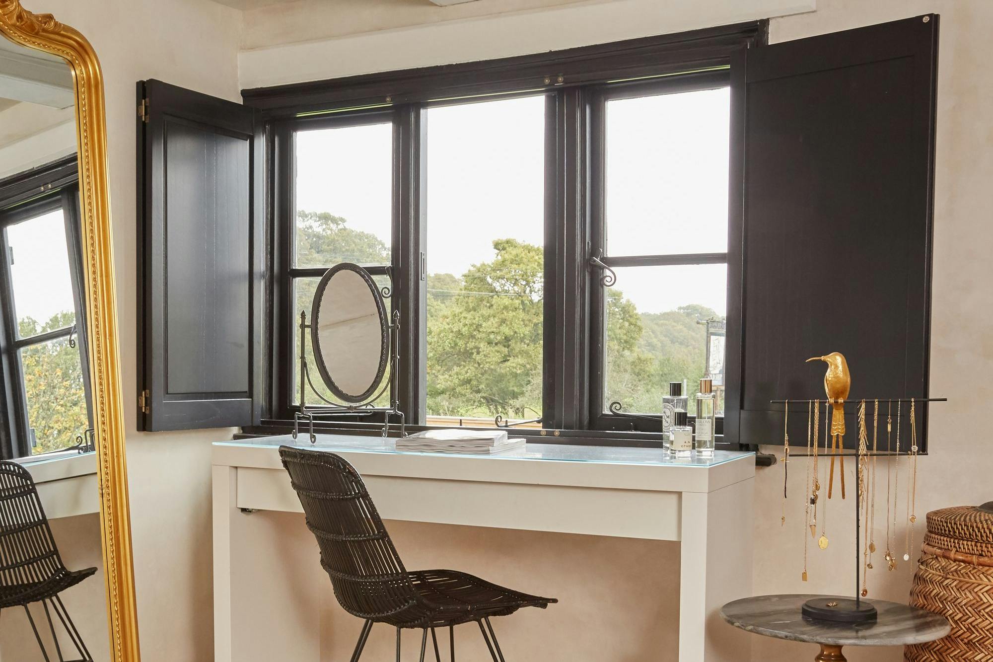 A rattan chair is positioned by a white vanity table with cosmetics, near black-framed windows overlooking greenery. A gold jewelry holder on a small table complements the cozy, elegant room.