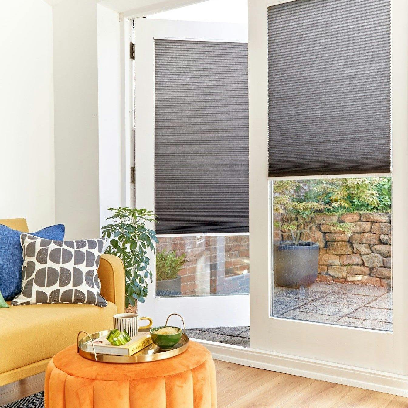 A cozy room features a mustard sofa with patterned cushions, next to a white door with dark blinds. Outside, a brick patio and stone wall are visible, enhancing the homely atmosphere.