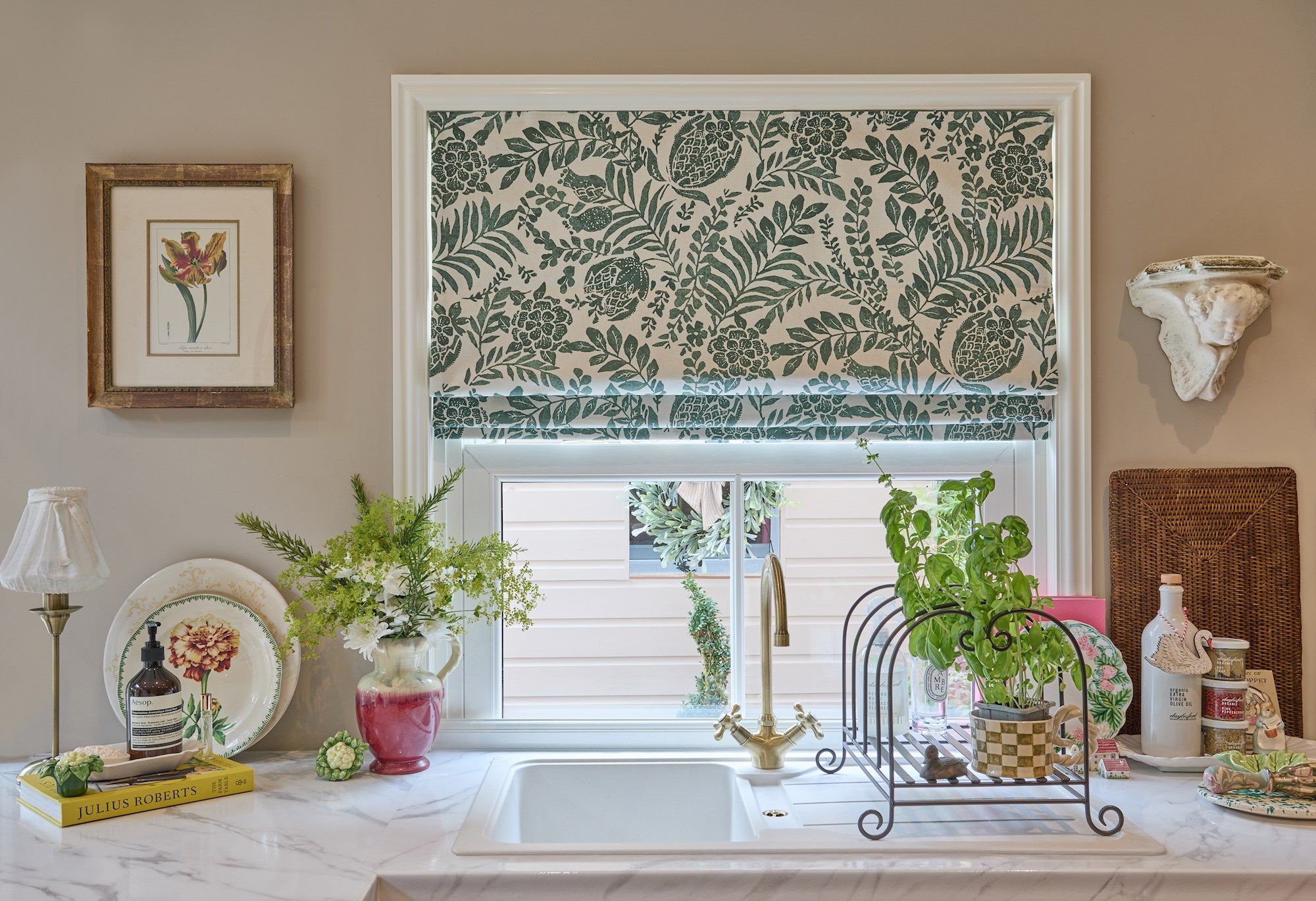 A kitchen sink with a gold faucet is surrounded by decorative plants and kitchenware. Above, floral-patterned blinds partially cover the window. Nearby, a lamp and framed flower art adorn the walls. Text includes "JULIUS ROBERTS."