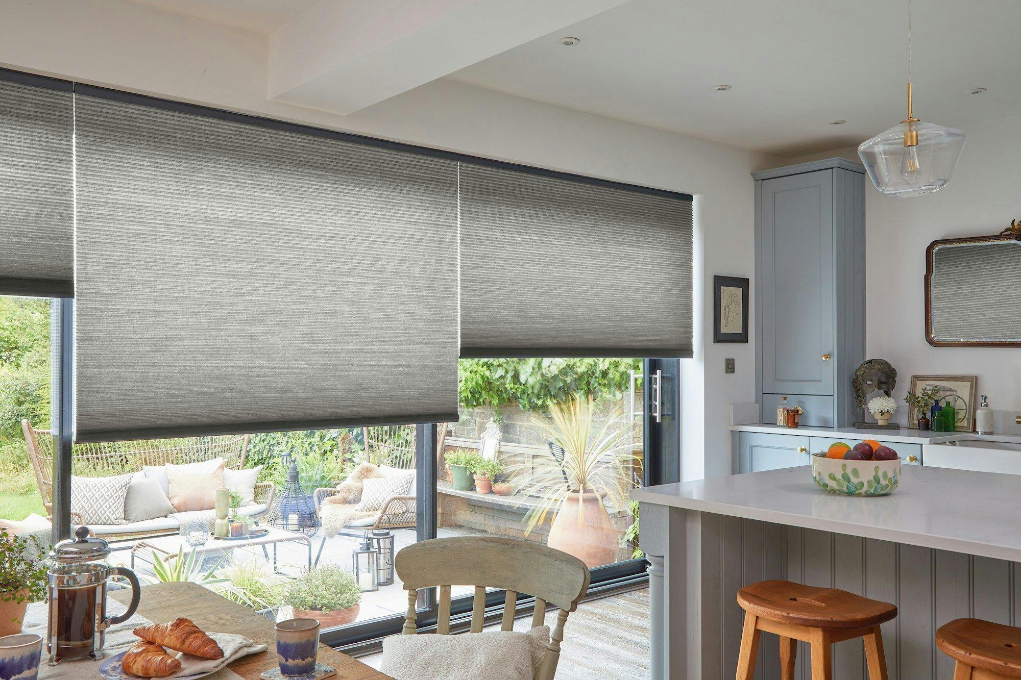 Gray blinds cover large windows overlooking a patio with plants and seating. A cozy kitchen features a counter with fruit, chairs, and decorative items, creating a warm atmosphere.