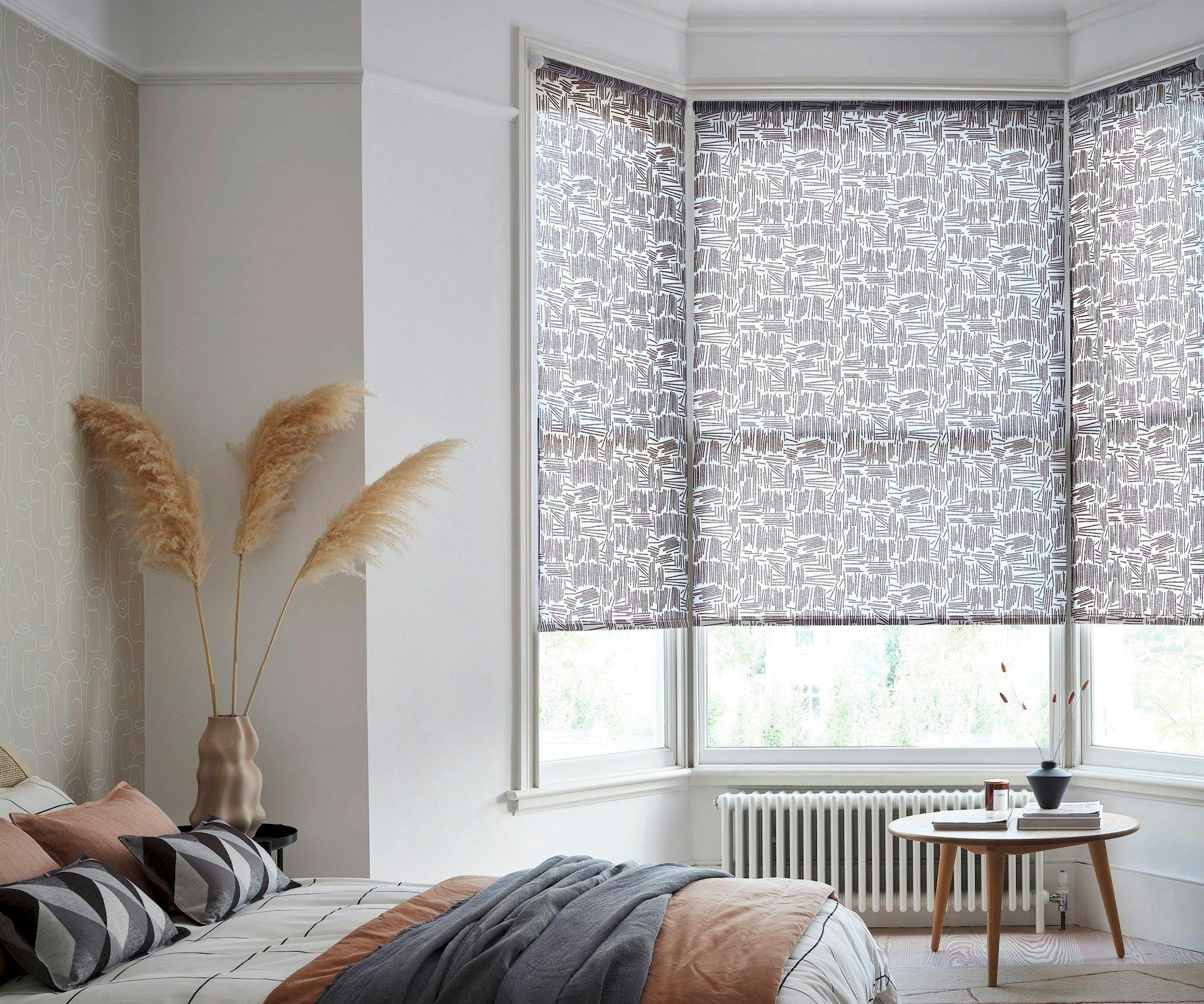 A bed with patterned pillows and blanket rests beside a vase with pampas grass in a bright room featuring a round table, radiators, and large windows with geometric blinds.