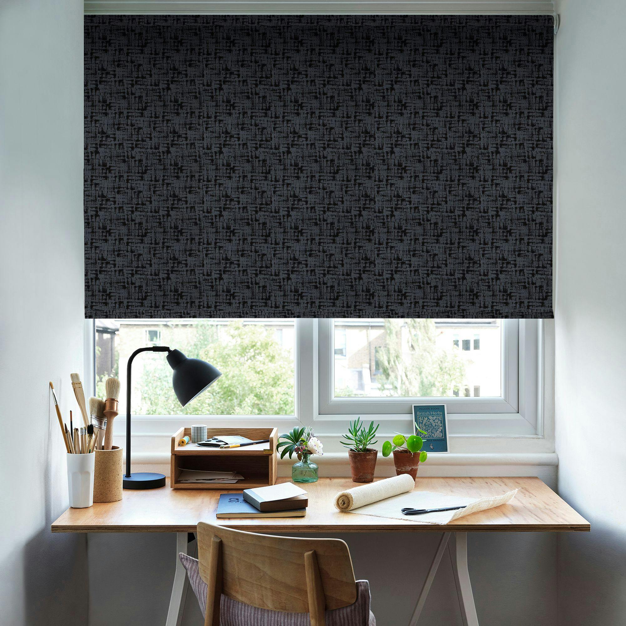 A wooden desk with various plants, books, and art supplies sits by a window. A dark, patterned roller blind covers the window, partially blocking the view of greenery outside.
