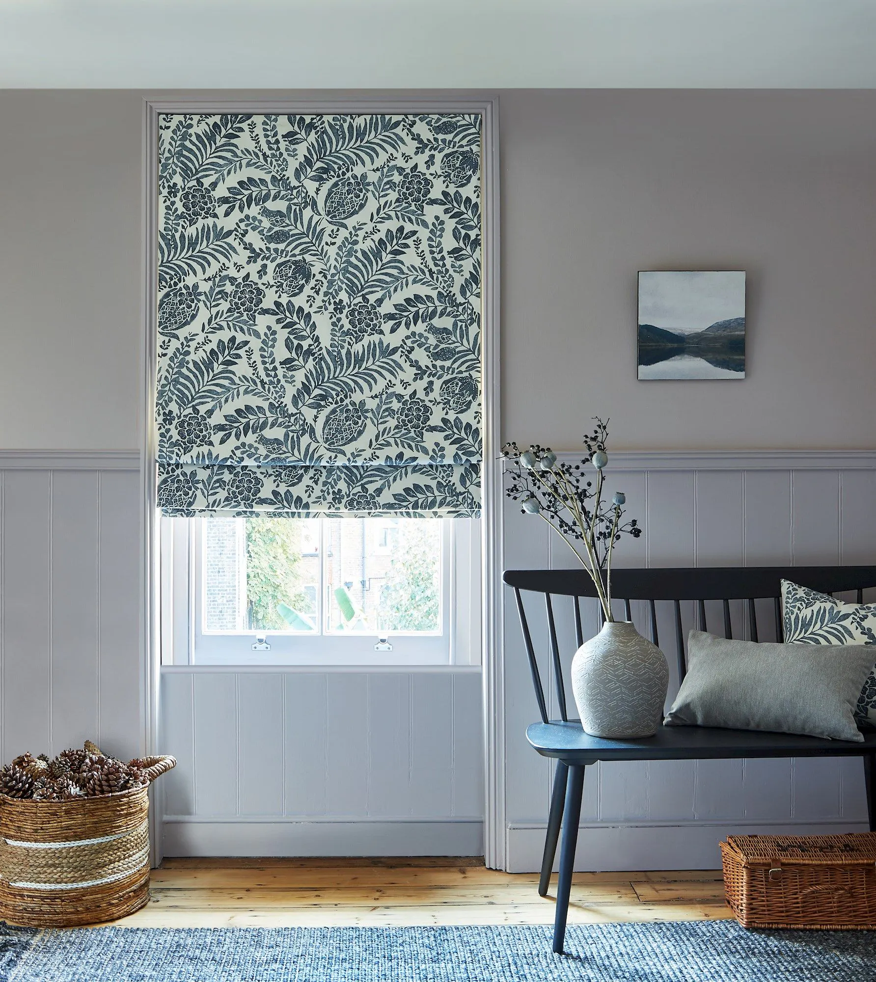 A floral-patterned roller blind covers a window in a cozy room. A bench with cushions and a vase of branches sits nearby on wooden flooring, beside a basket of pinecones.