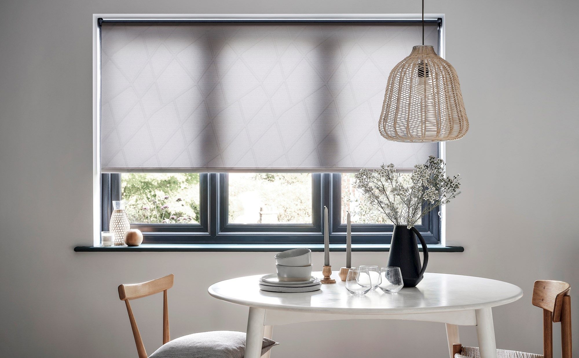 A round white table holds stacked plates, cups, and glasses, with a black vase and candles. A woven pendant light hangs above. Roller blind decorates the window in a minimalist room.